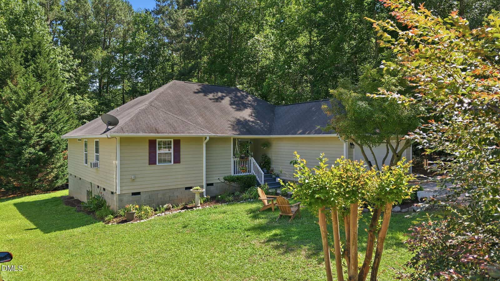 A single-story yellow house with a dark roof sits amid green trees and lawn, with a covered front porch, landscaping, and wooden chairs on the grassy yard under a sunny sky.