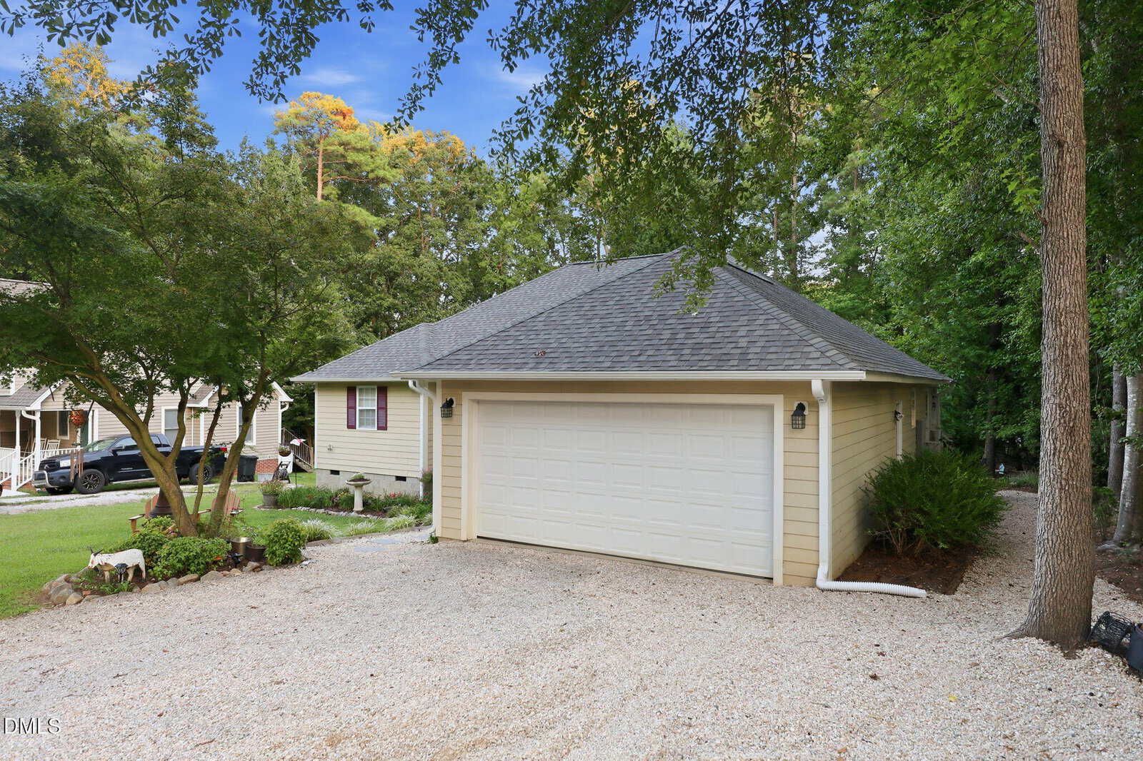 A light yellow house with a gray roof and white double garage, surrounded by trees and greenery, with a gravel driveway in front.