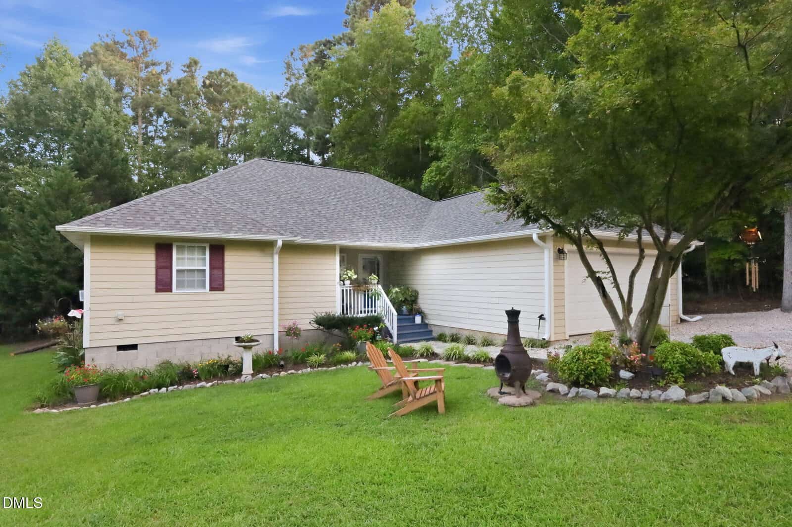 A single-story beige house with a gray roof, front porch, flower beds, green lawn, wooden chairs, and a chiminea. Surrounded by trees and a gravel driveway on the right.