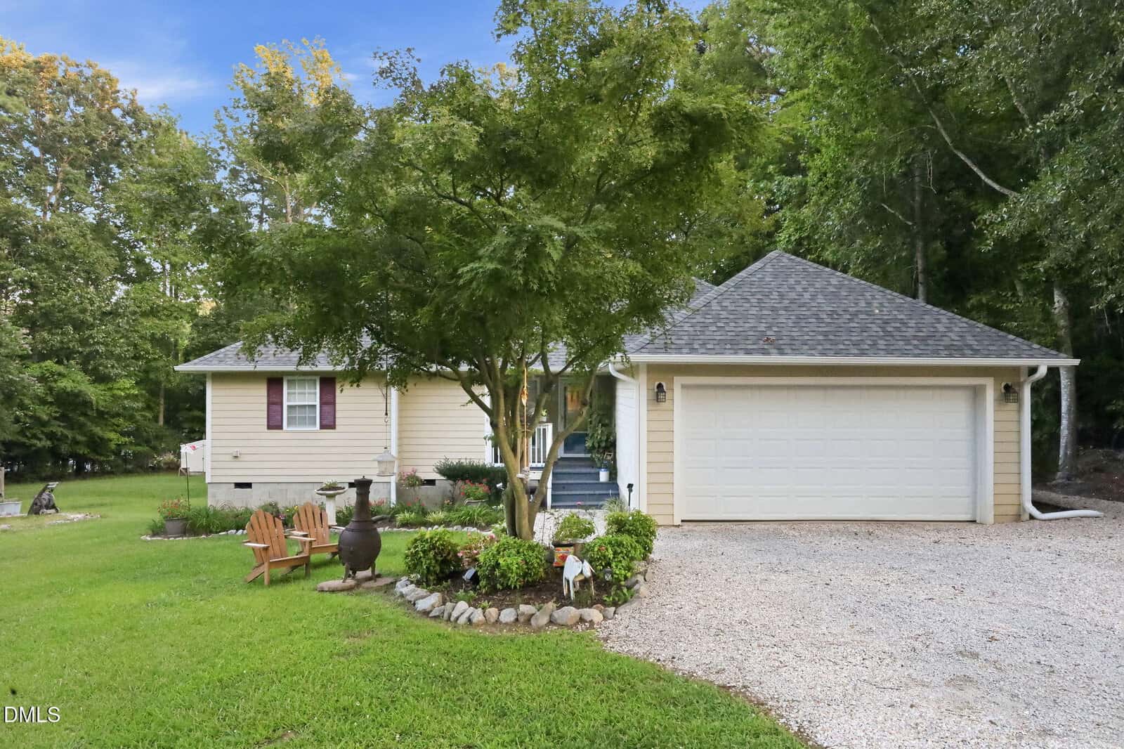 Charming single-story beige house at 106 Yuma Drive, Louisburg, featuring a two-car garage and surrounded by green lawn and trees. A gravel driveway leads to the garage; a small garden and Adirondack chairs sit in the front yard. Blue sky is visible above.