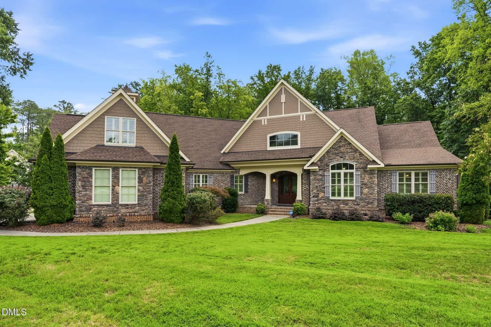 A large, two-story house with stone and wood siding, tall peaked roofs, arched windows, green lawn, and shrubs, set against a backdrop of trees under a blue sky at 1106 Dovefield Lane in Youngsville.