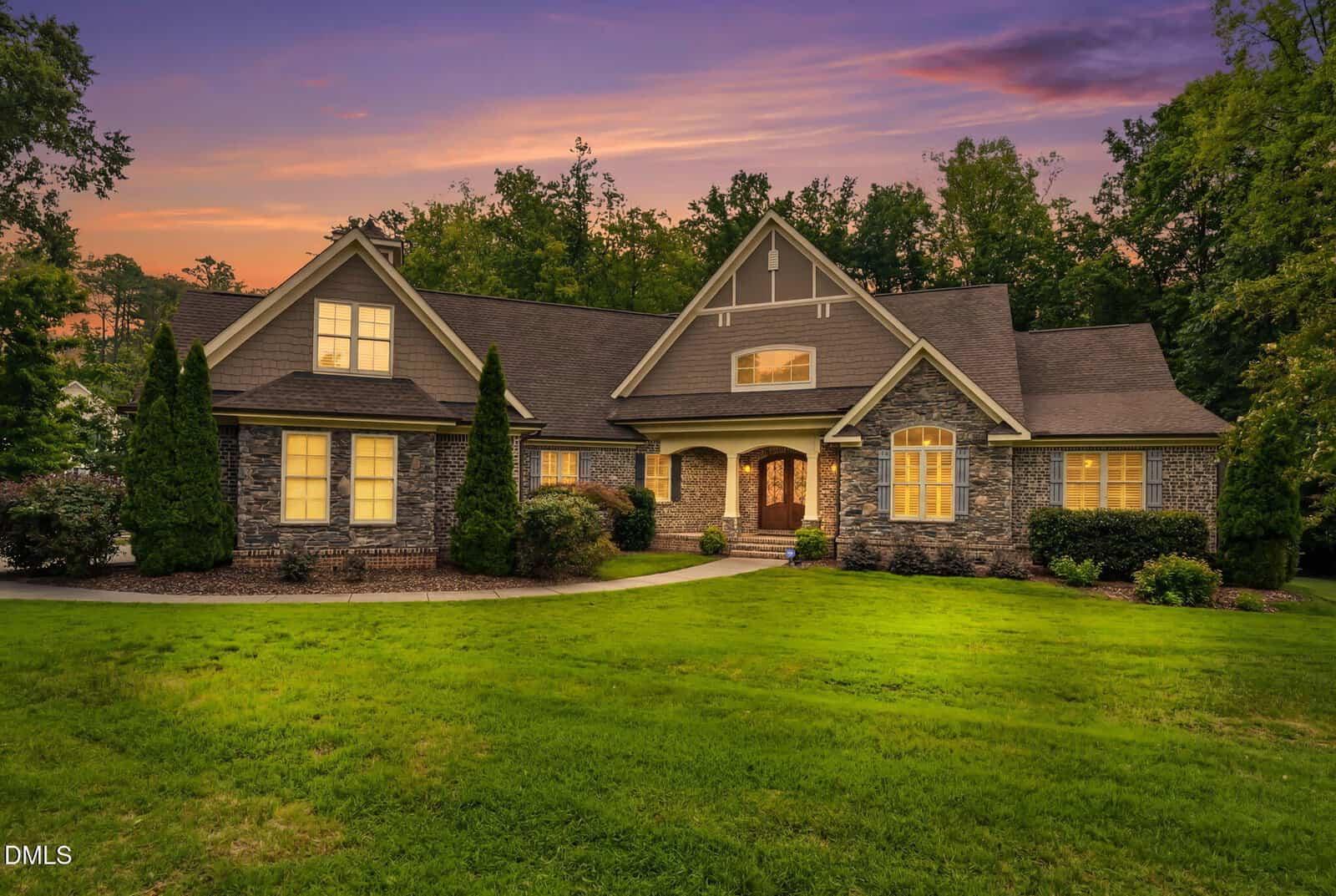 Stone and brick house with large windows, peaked roof, and manicured lawn, set against a colorful sunset sky with trees in the background.