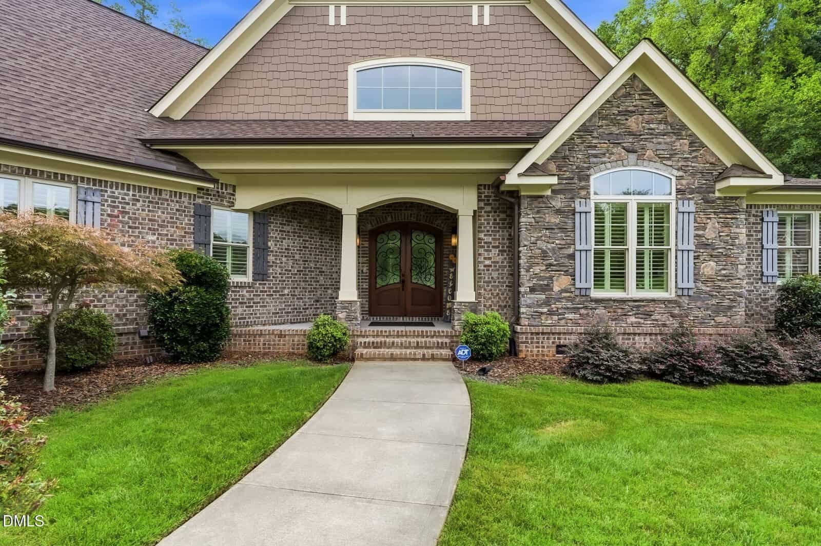 Curved sidewalk leads to double wooden front doors of a suburban home with brick and stone exterior, arched entryway, manicured lawn, shrubs, and shuttered windows.