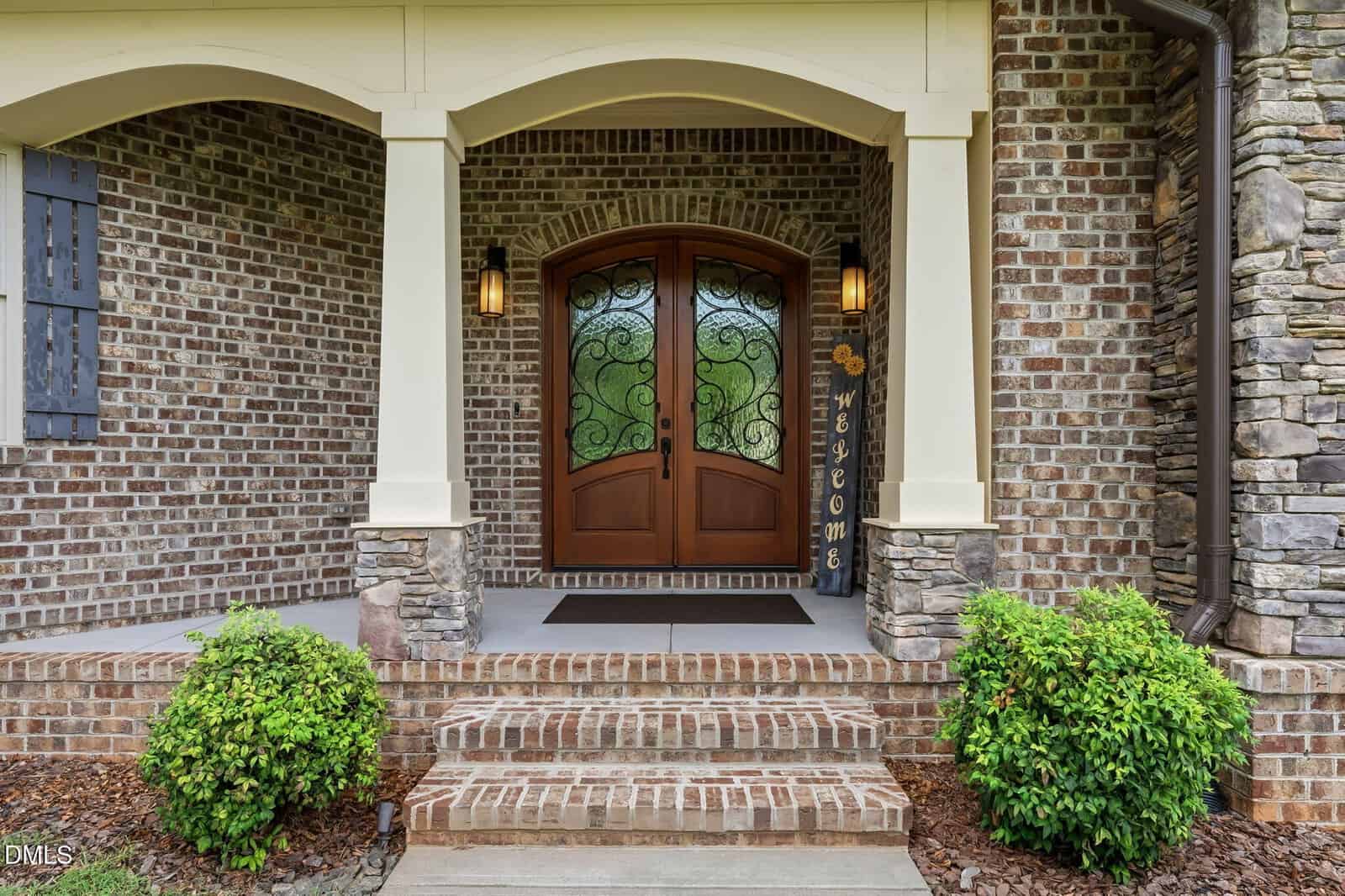Brick house entrance with double wooden doors featuring decorative glass, flanked by two lantern lights, a "Welcome" vertical sign, stone accents, steps, and two green bushes on either side.