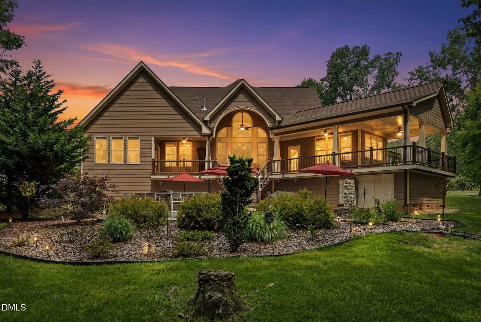 A large, brown house with a covered porch and patio, glowing warmly at sunset, is surrounded by landscaped gardens, trees, and outdoor seating areas with red umbrellas.