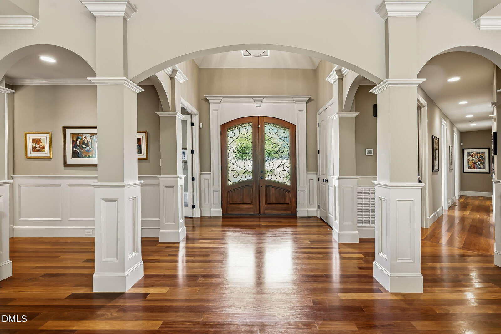 Elegant home entryway with polished wood floors, arched double doors featuring ornate ironwork, white columns, neutral walls, and framed artwork along the hallway. Bright lighting creates a welcoming atmosphere.