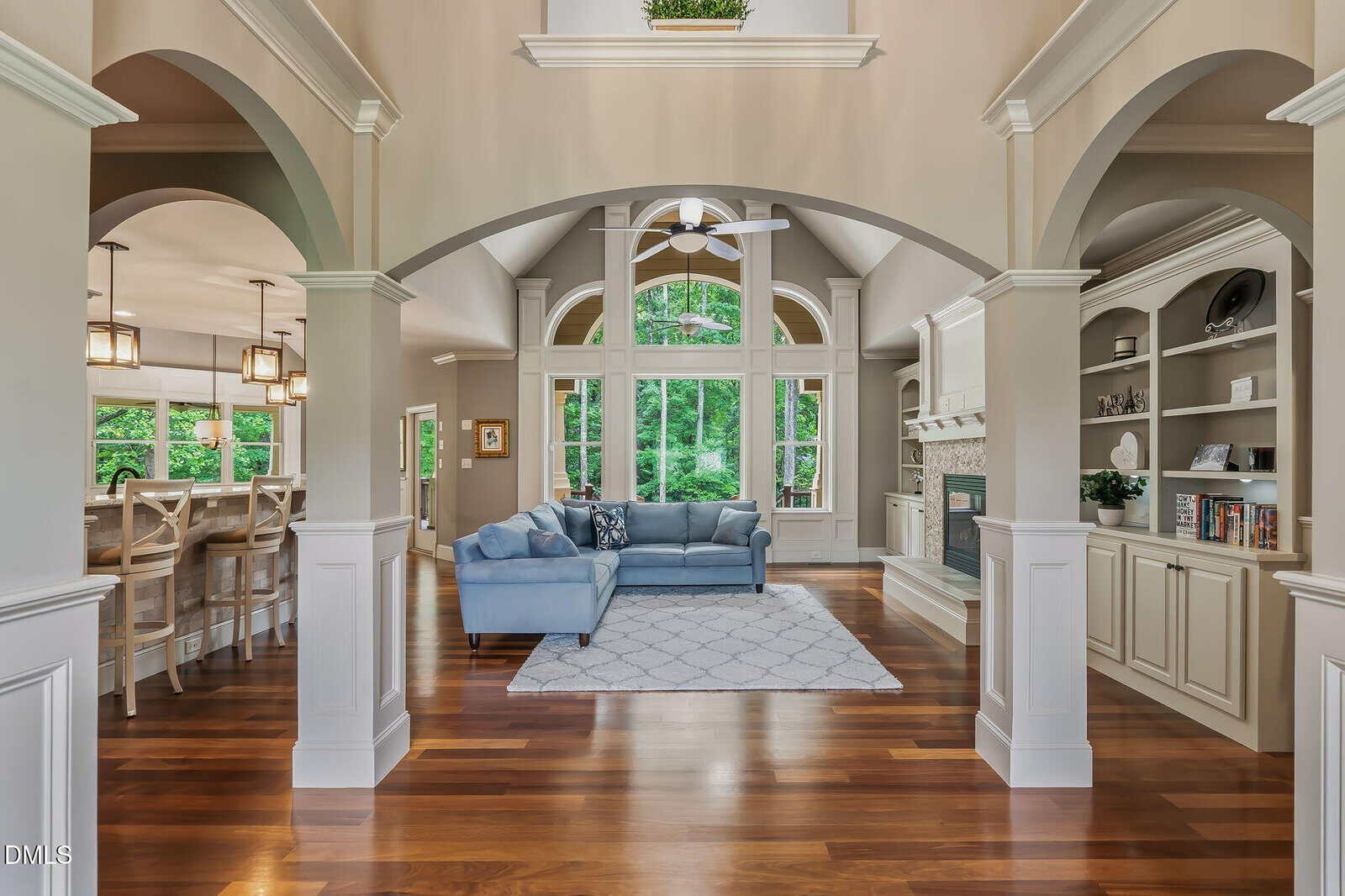 Open living room with high ceilings, large windows, blue sectional sofa, built-in shelves, fireplace, and wooden floors. Dining area with barstools visible to the left. Neutral wall colors and lots of natural light.