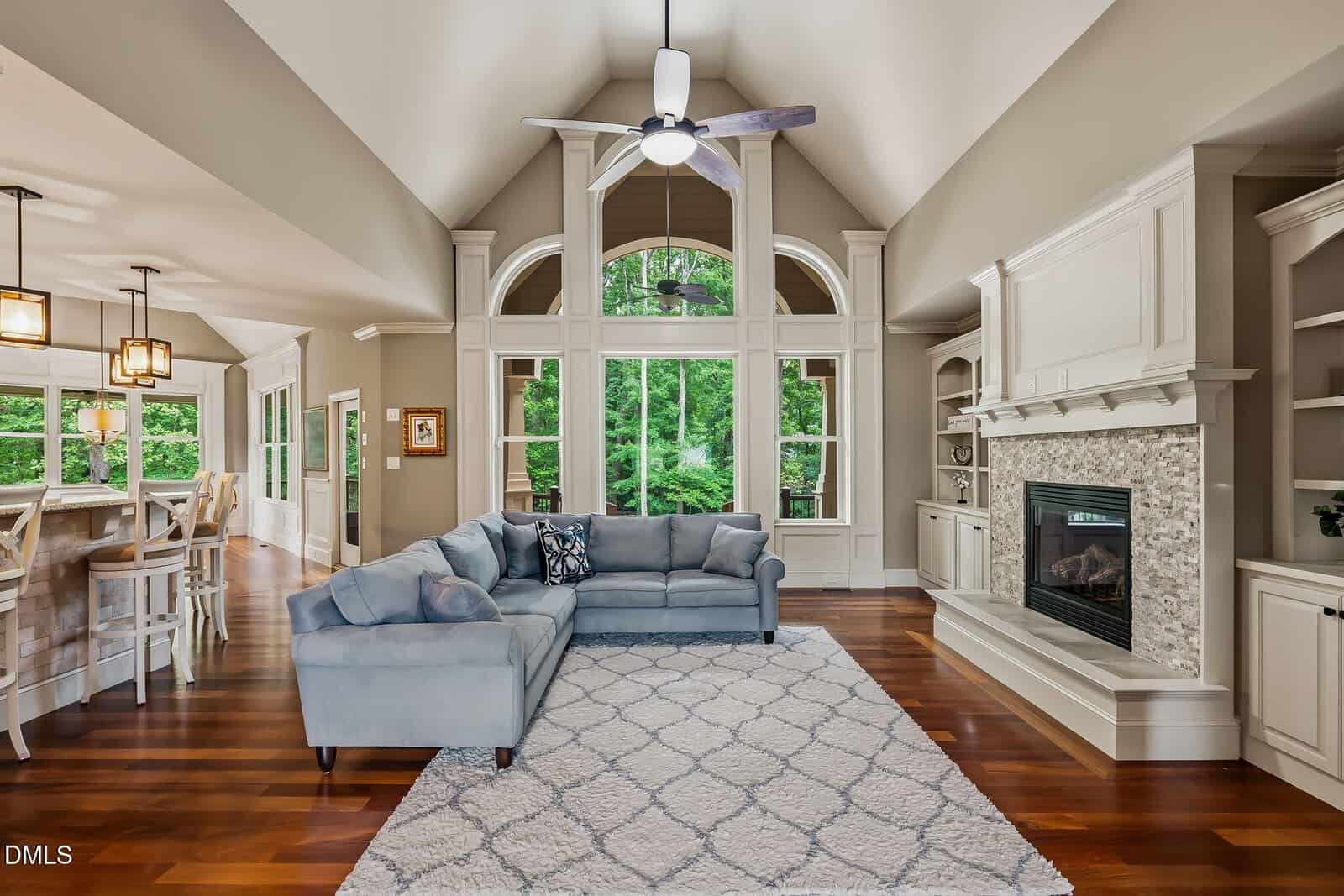 Spacious living room with hardwood floors, gray couches, a patterned rug, a white fireplace, built-in shelves, large windows, and a ceiling fan. Open layout connects to a kitchen with bar seating.