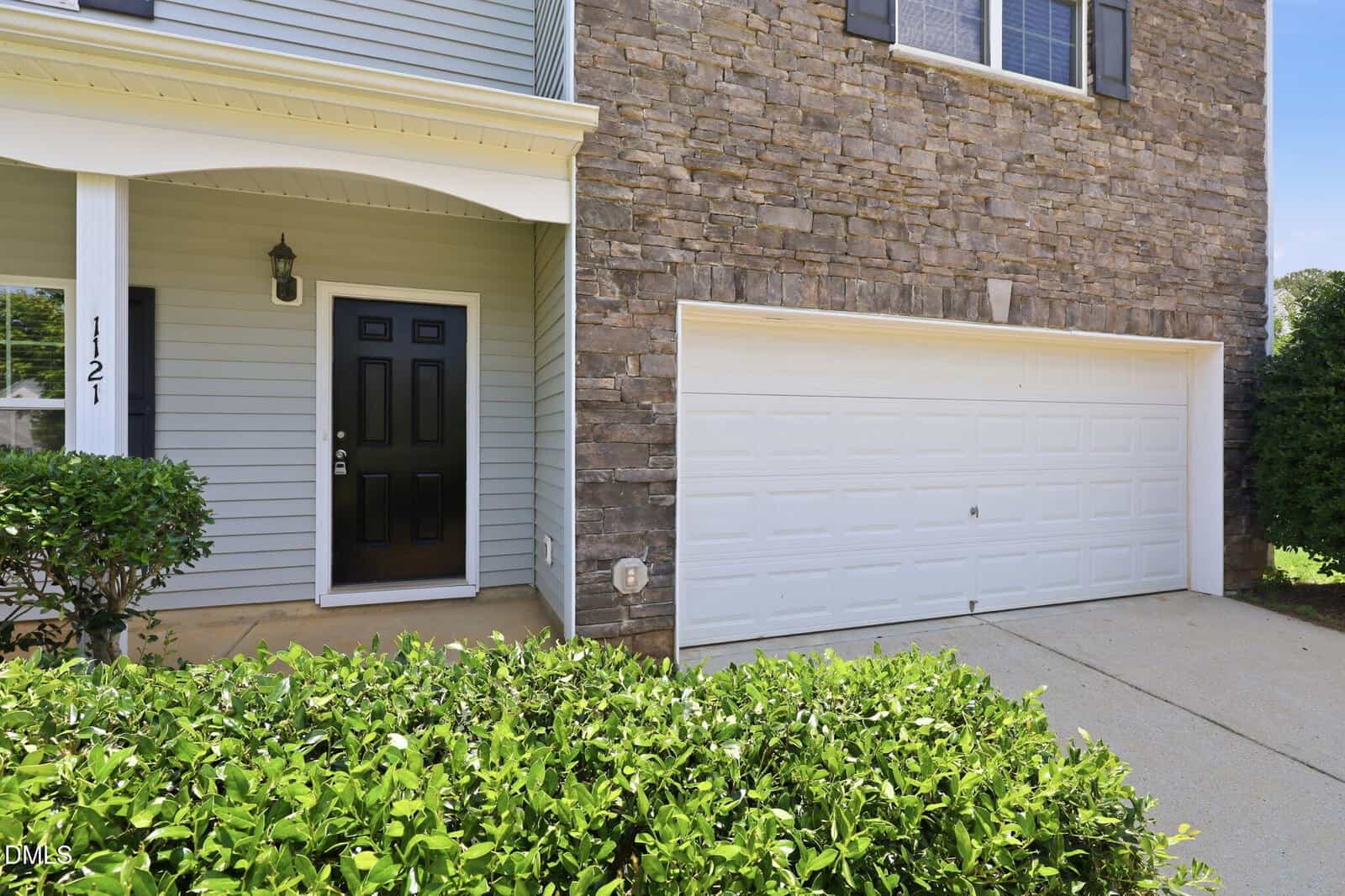 Front exterior of a house with a black door, light gray siding, stone accent wall, white double garage door, and green hedges in the foreground.