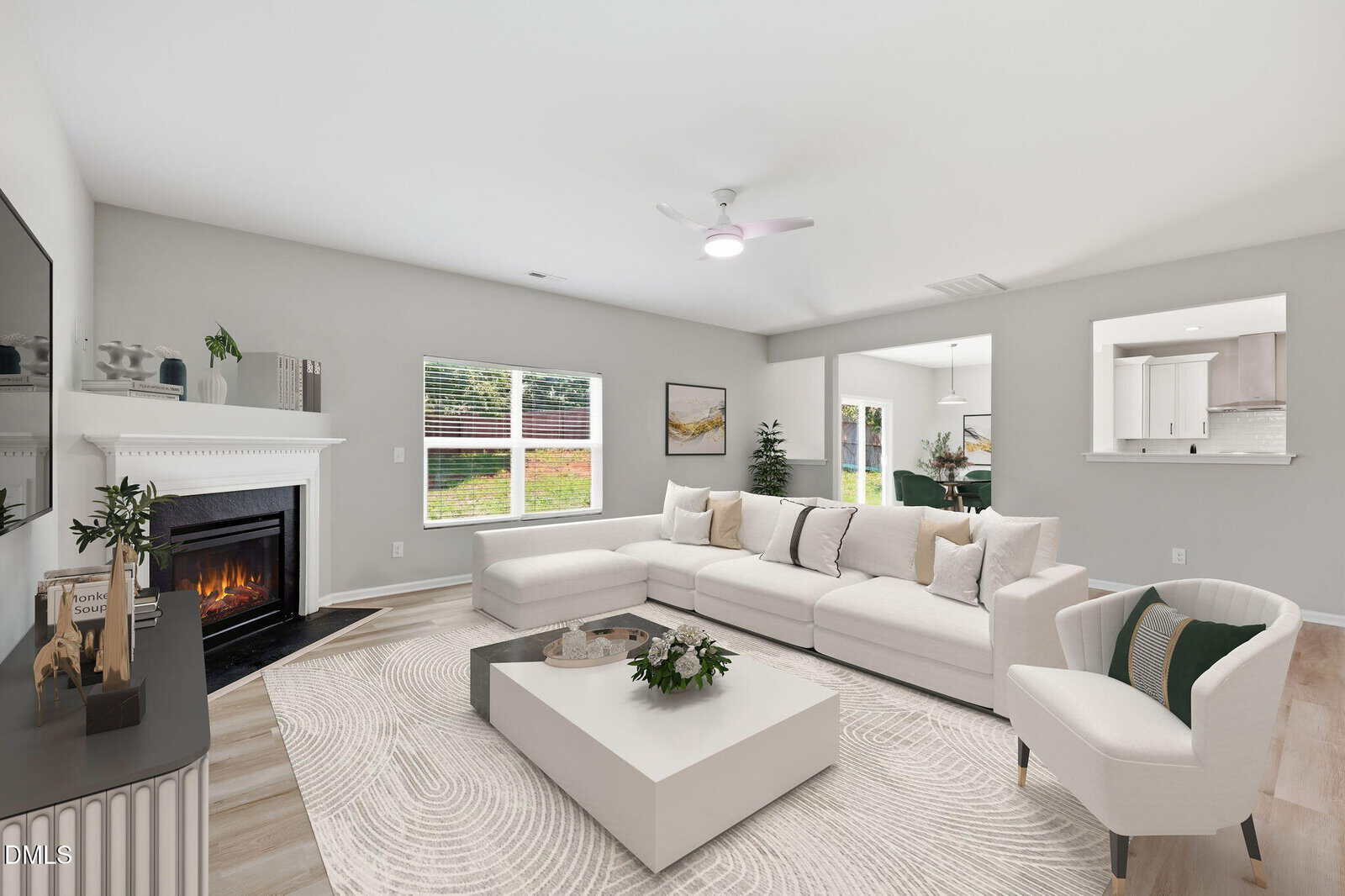 Bright living room with a white sectional sofa, armchair, fireplace, coffee table, and decor. Large window shows green lawn. Open plan connects to kitchen and dining area with light wood floors and neutral walls.