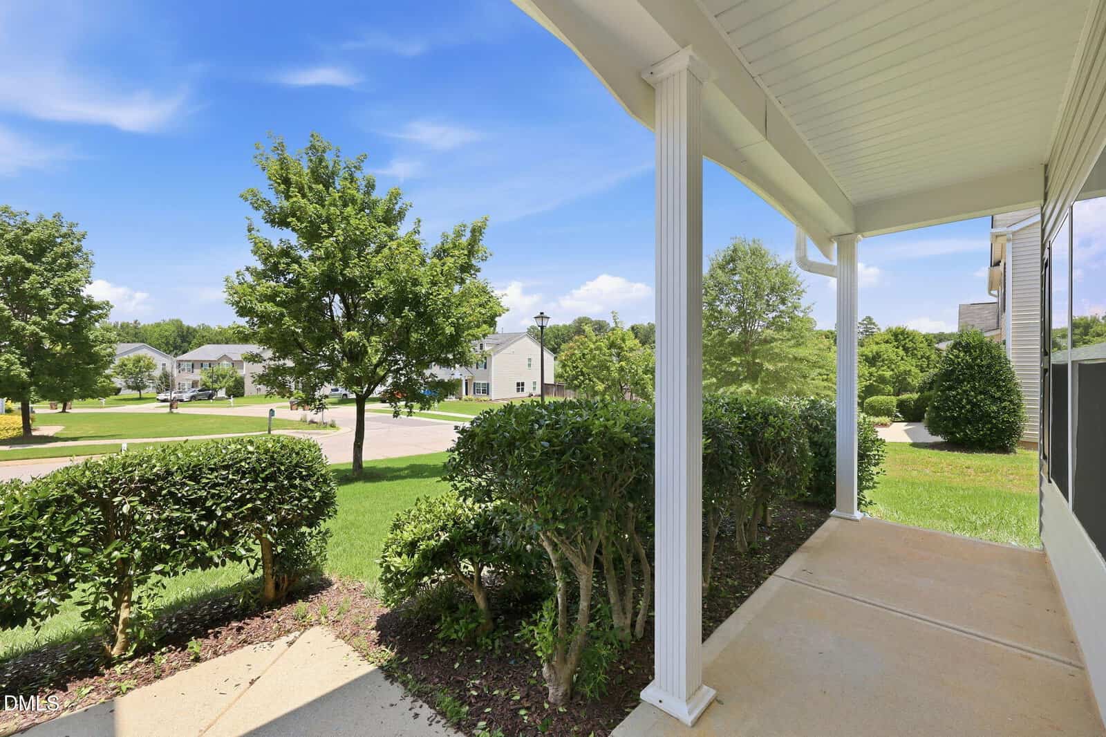 View from a covered porch with white columns, looking out onto a green lawn, trimmed bushes, trees, and a quiet suburban street with houses under a blue sky with some clouds.