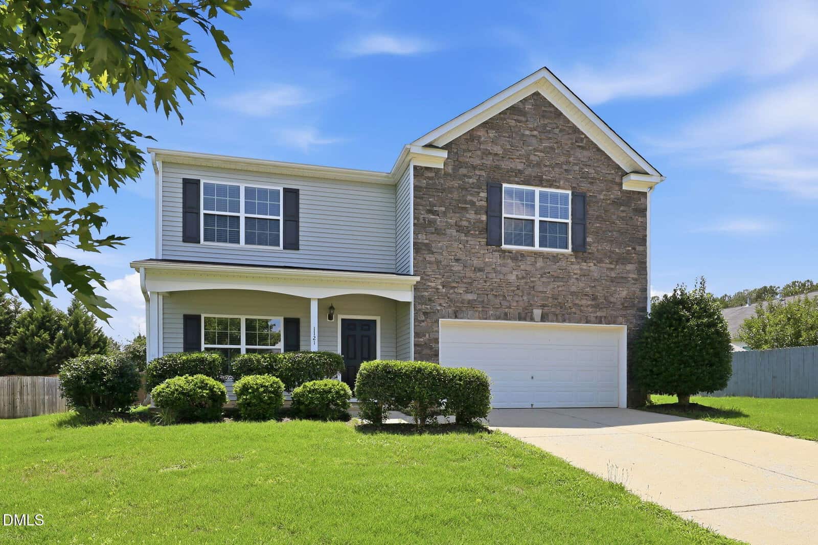 Two-story suburban house at 1121 Crendall Way, Wake Forest, with a stone and siding exterior, black shutters, white trim, and a double garage. Green lawn, bushes, and a tree in the yard complement the clear blue sky overhead.