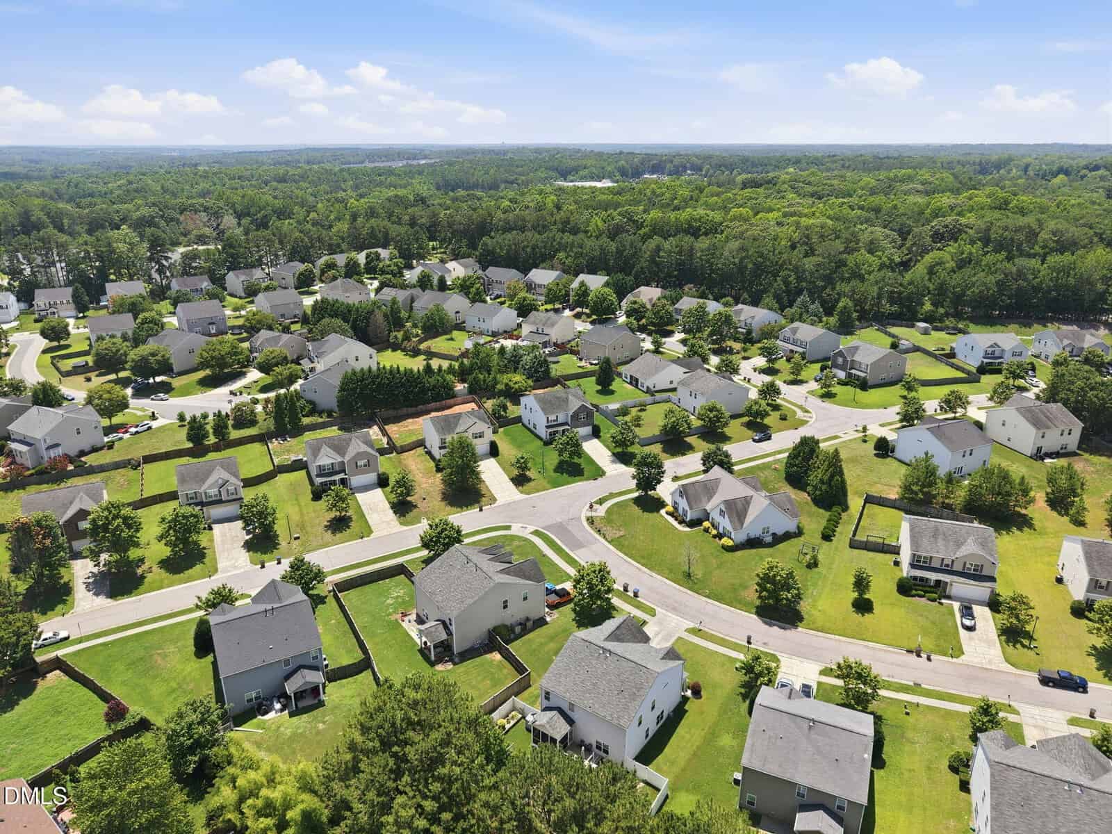 Aerial view of a suburban neighborhood with rows of single-family homes, green lawns, curved streets, and trees under a blue sky with scattered clouds.