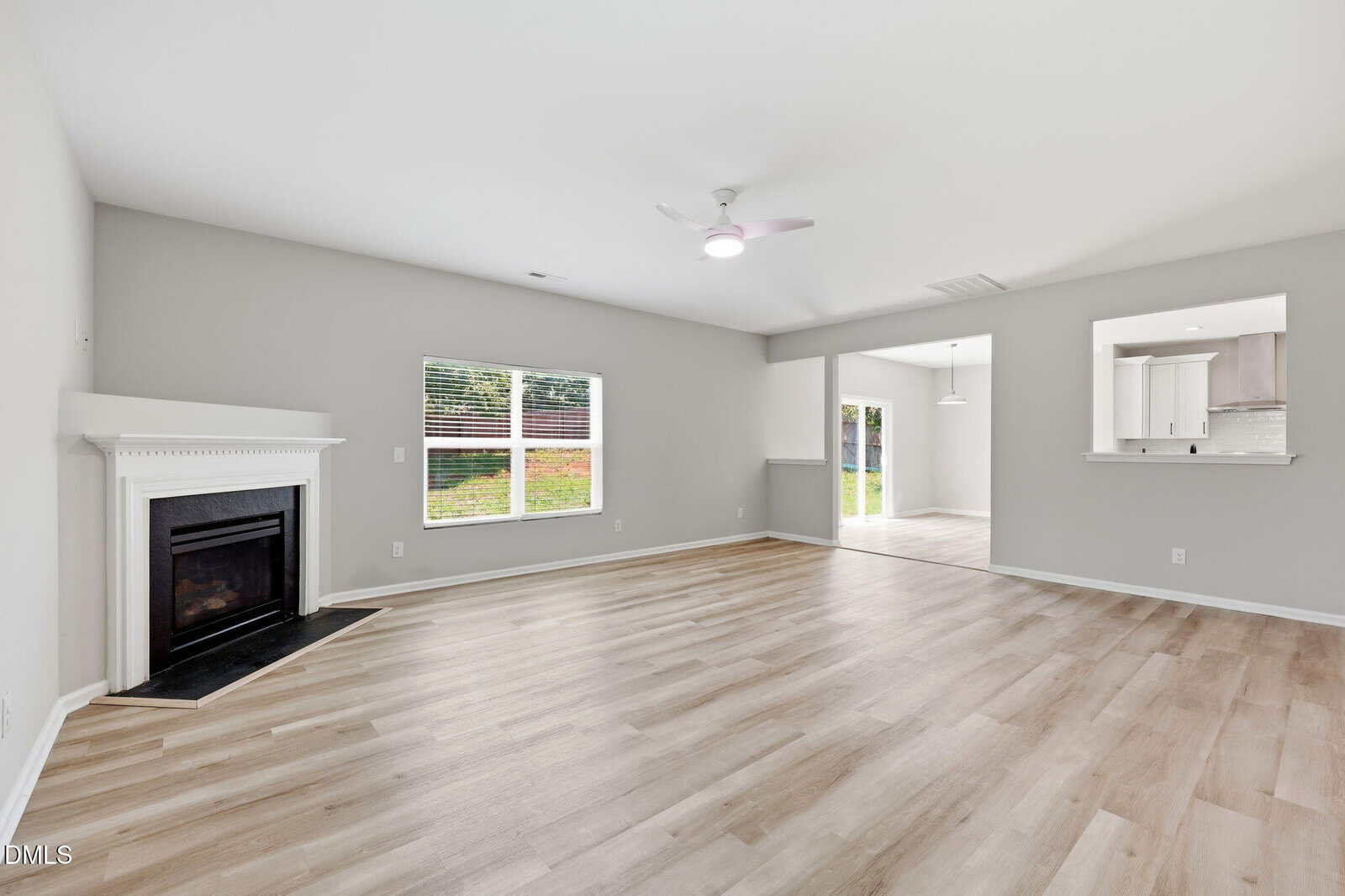 Bright, spacious living room with light wood flooring, a white fireplace in the corner, large window, ceiling fan, and open access to dining area and kitchen with neutral walls.