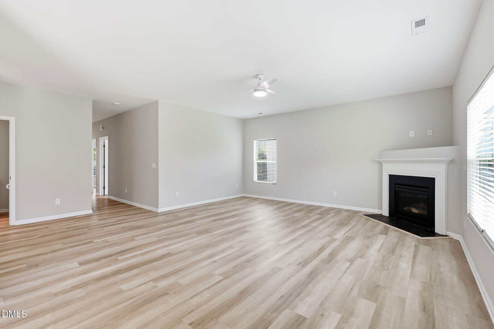 Spacious, empty living room with light wood flooring, white walls, large windows, a ceiling fan, and a corner fireplace with a white mantel. Doorways lead to other rooms. Natural light fills the space.