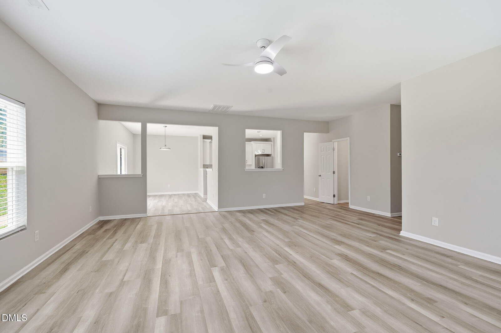 Spacious, empty living area with light wood floors, neutral walls, ceiling fan, and large windows. Open layout leads to dining area and kitchen with modern fixtures and natural light.