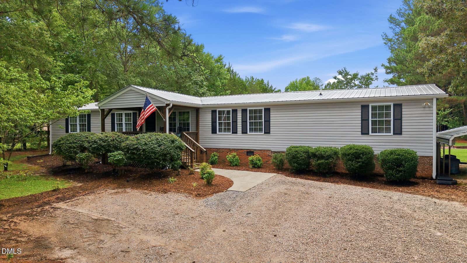 Single-story gray manufactured home with white trim, black shutters, and a metal roof at 104 Pineview Drive, Louisburg. An American flag hangs by the front porch. The yard has green bushes, trees, and a gravel driveway under a blue sky.