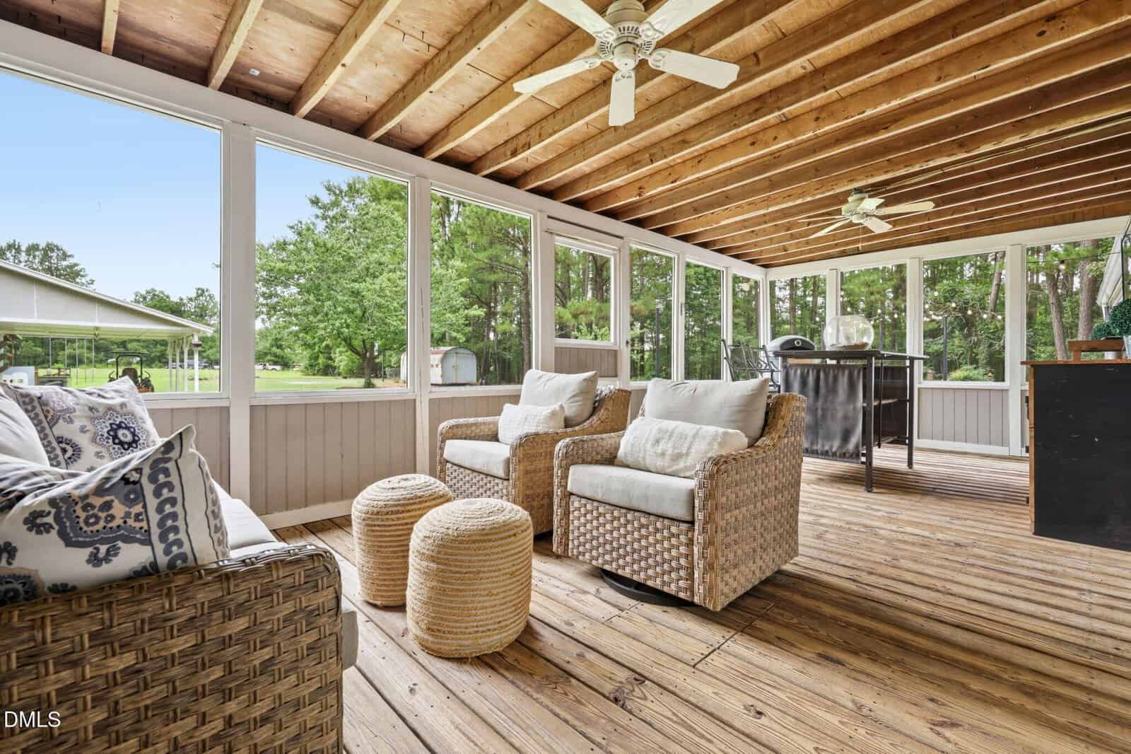 Screened-in porch with a wood floor and exposed beams, featuring wicker furniture with cushions and pillows, two poufs, ceiling fans, and large windows overlooking a green yard.