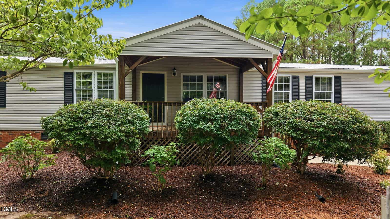 A single-story house with beige siding, black shutters, and a covered front porch, surrounded by trimmed bushes and mulch. An American flag hangs by the porch, and trees are visible in the background.