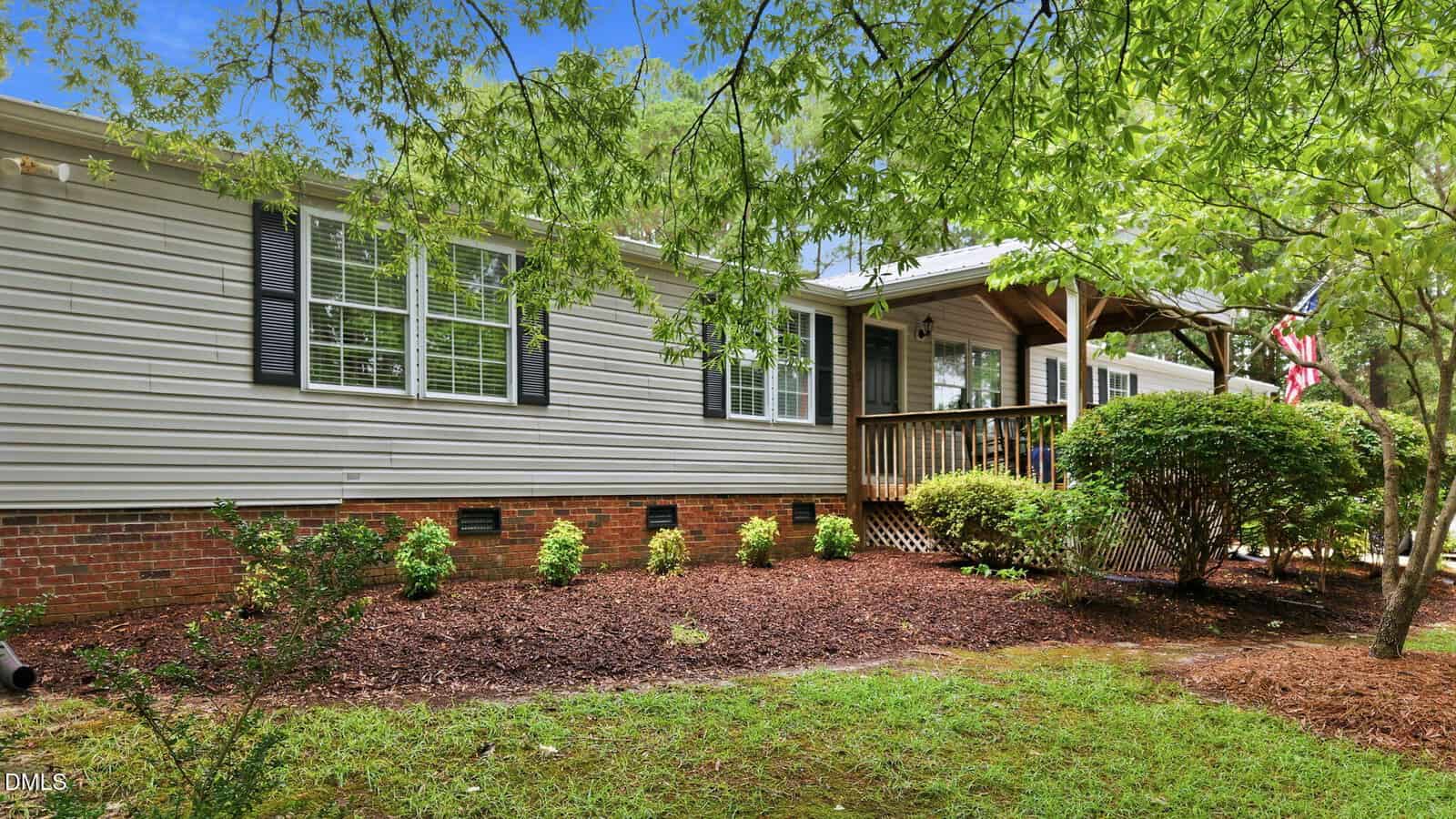 Single-story house with light-colored siding, black shutters, a brick foundation, and a covered front porch, surrounded by green shrubs, trees, and a well-kept lawn.