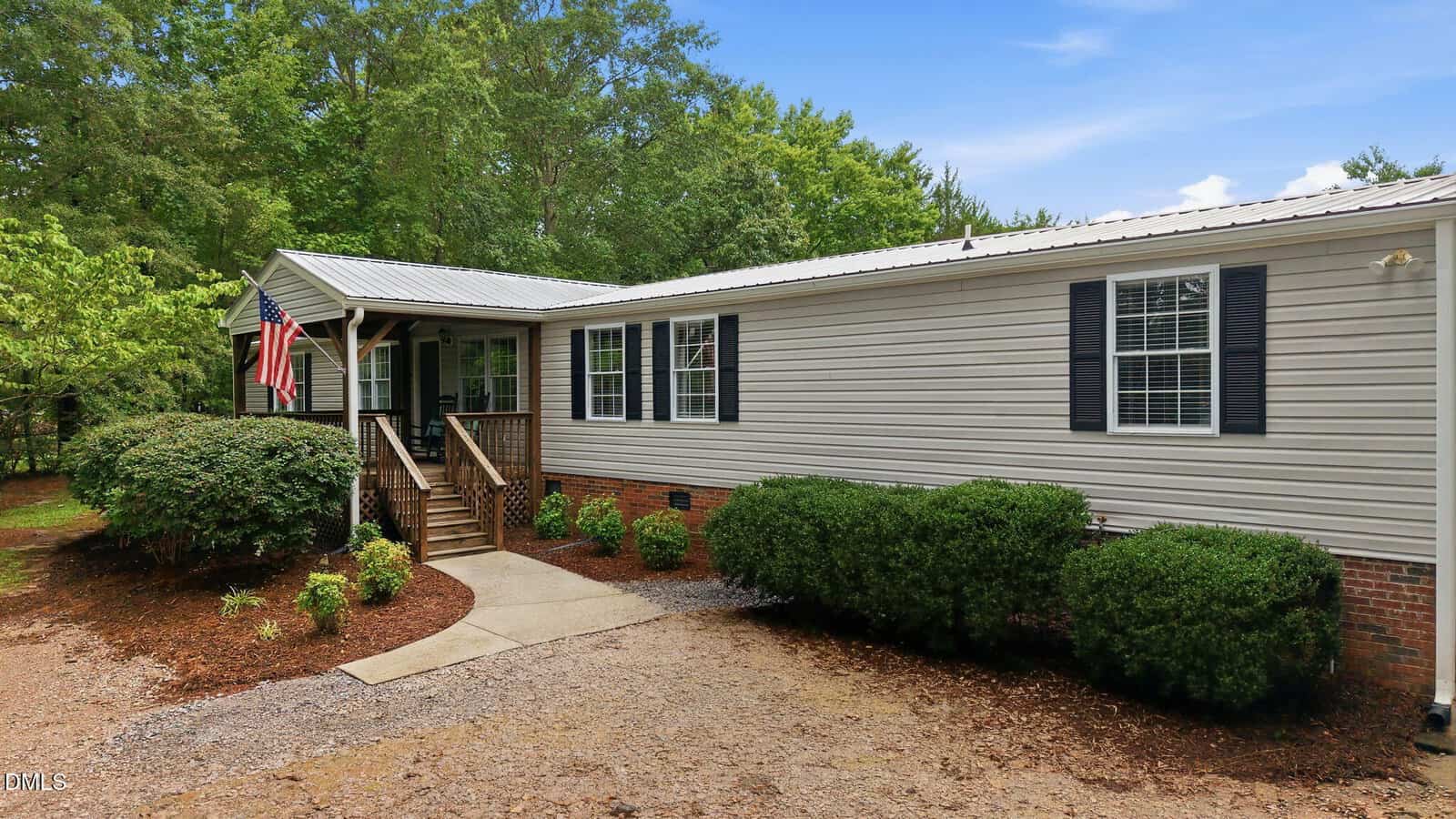 Single-story beige manufactured home with a front porch, black shutters, and a metal roof. An American flag hangs by the door. Shrubs and trees surround the house under a blue sky.
