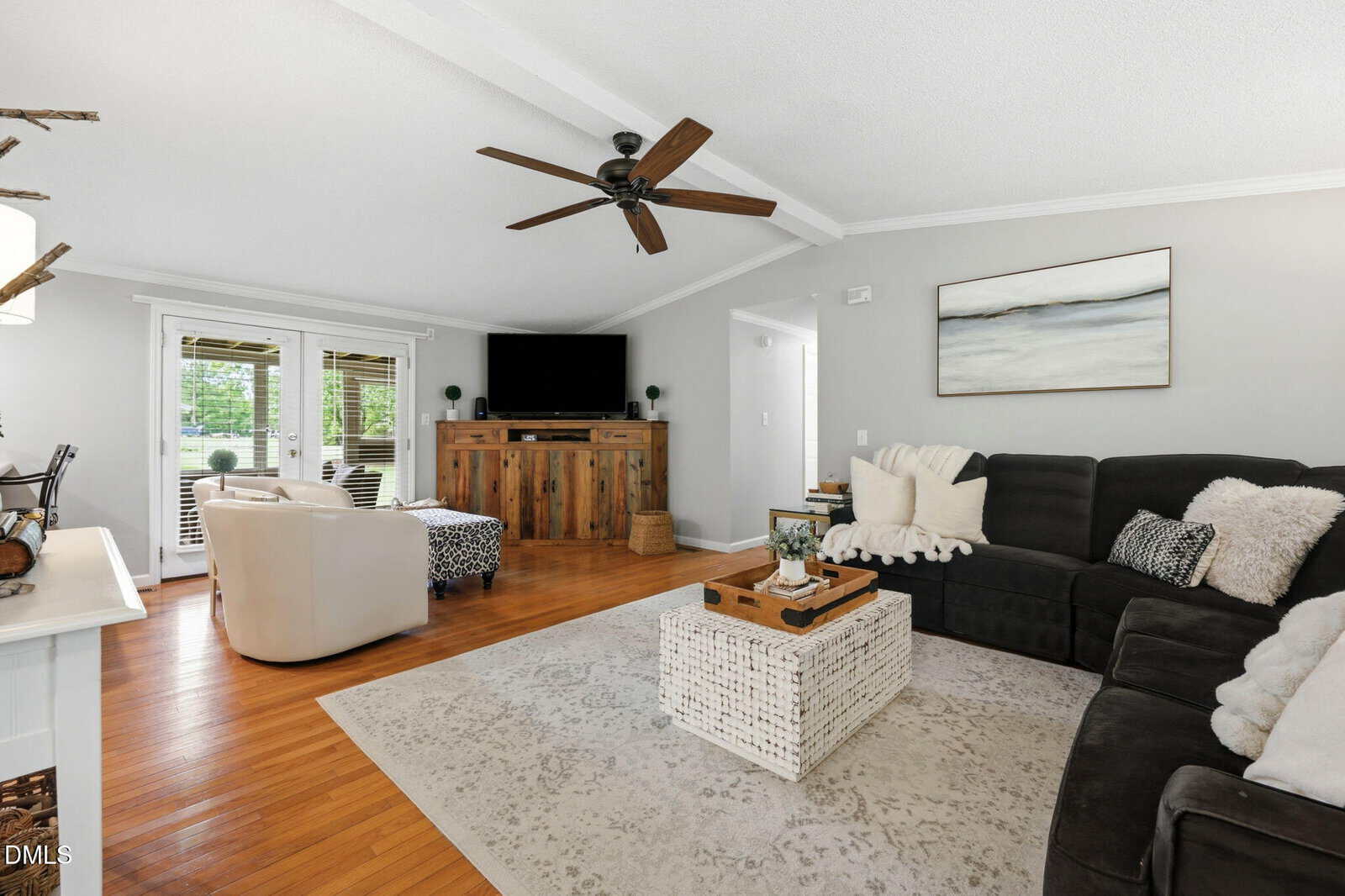 Bright living room with wood floor, ceiling fan, black sectional sofa, white armchair, TV on a rustic console, large area rug, and a coffee table; French doors let in natural light.