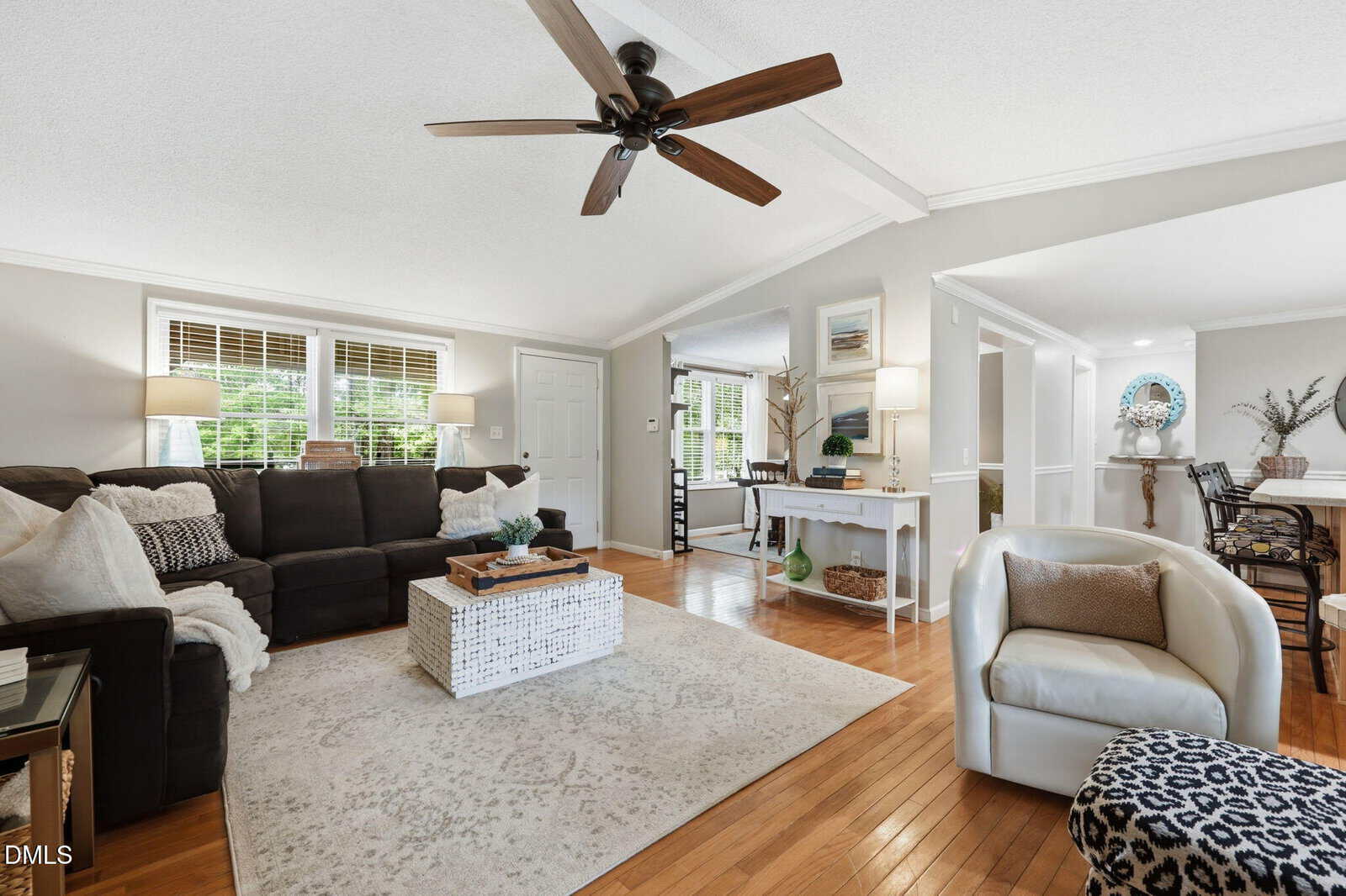 Bright living room with a sectional sofa, coffee table, area rug, ceiling fan, and hardwood floors. Open layout leads to dining area and entryway. White walls, large windows, and stylish decor create a welcoming atmosphere.