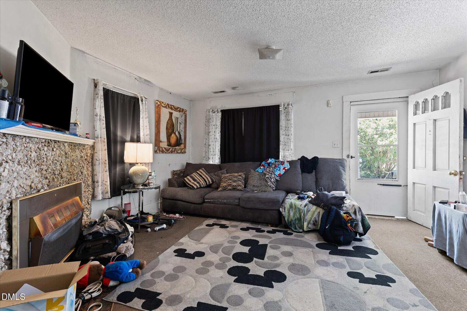 A cluttered living room with a gray sectional couch, patterned rug, TV above a fireplace, open door, scattered clothes, and various household items around the space.