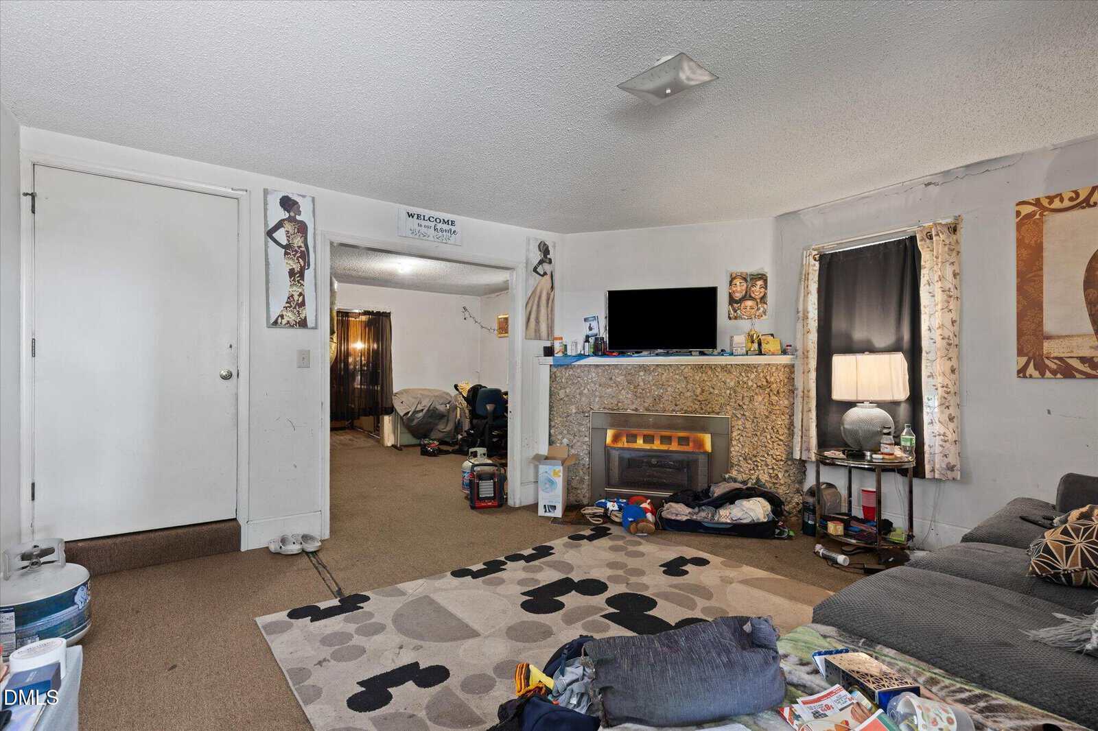 Living room with a couch, TV above an electric fireplace, various household items scattered around, and art on the walls. Doorway and window with curtains are also visible. The room has a lived-in, cluttered appearance.