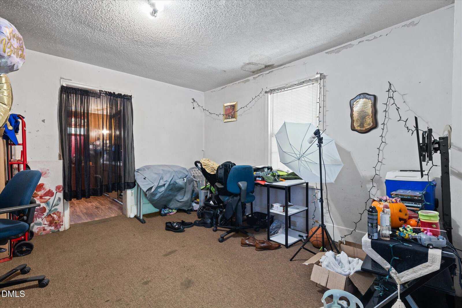 A cluttered home office with string lights, open storage shelves, two office chairs, photography umbrella, scattered items on the floor, and a desk with computer monitors. Light enters from a window with blinds.