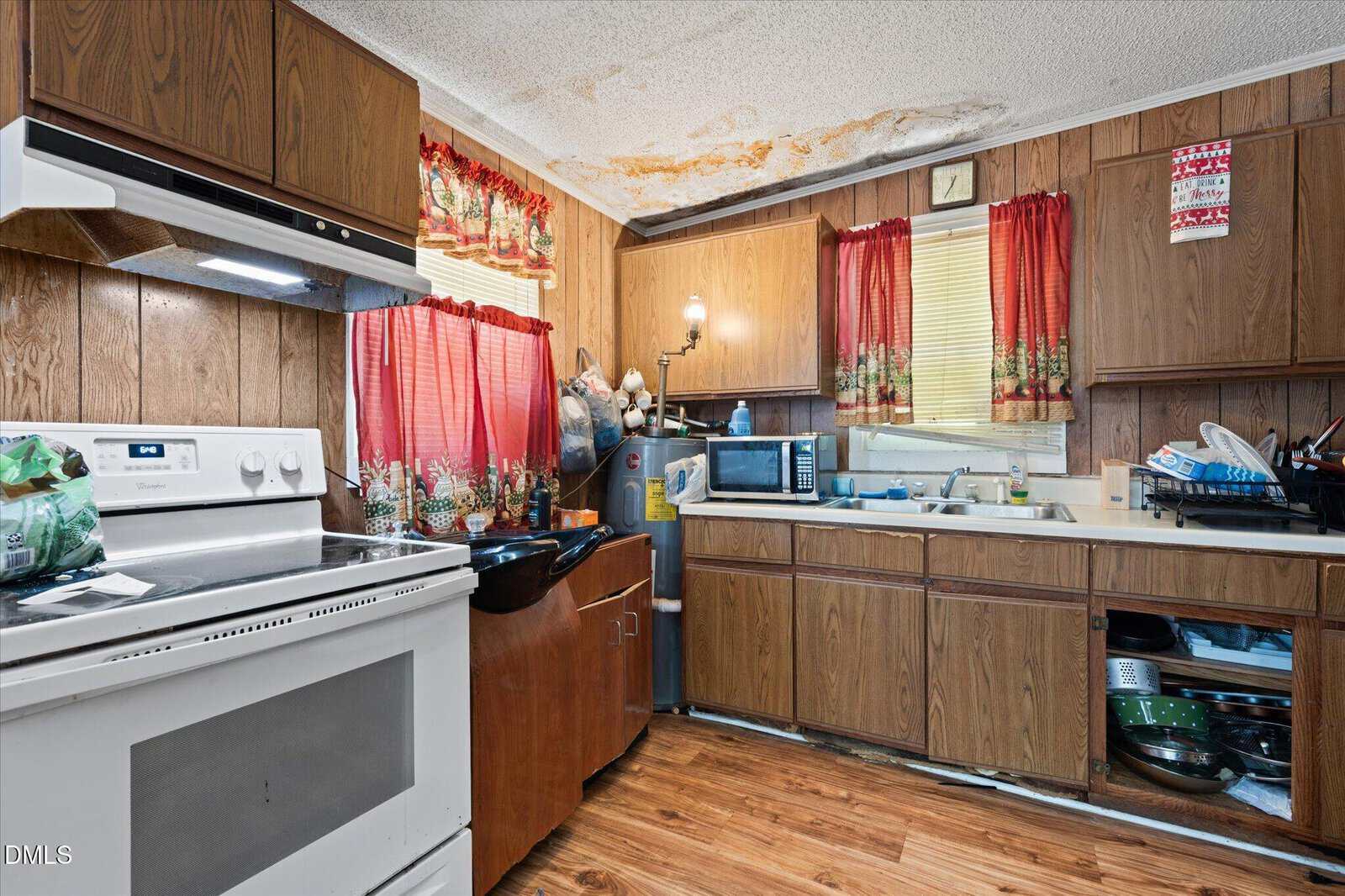 A cluttered kitchen with wood-paneled cabinets, red curtains, stove, sink, microwave, and dishes. The ceiling shows visible water damage and mold stains above the sink area.