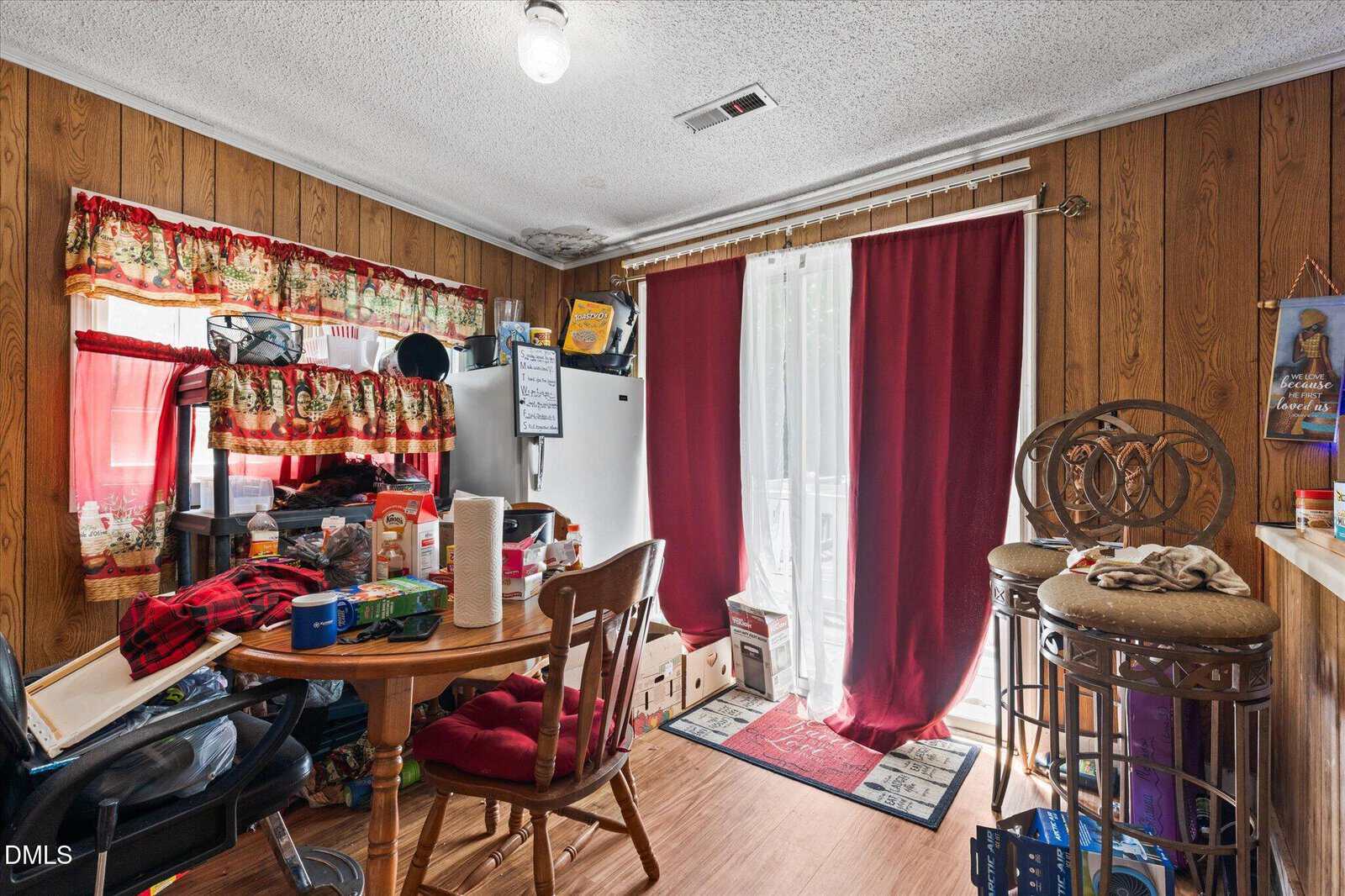 A cluttered dining area with wood-paneled walls, a round table with various items, a white fridge, red curtains over sliding doors, and a high chair. Light comes in through the curtains and a small window.