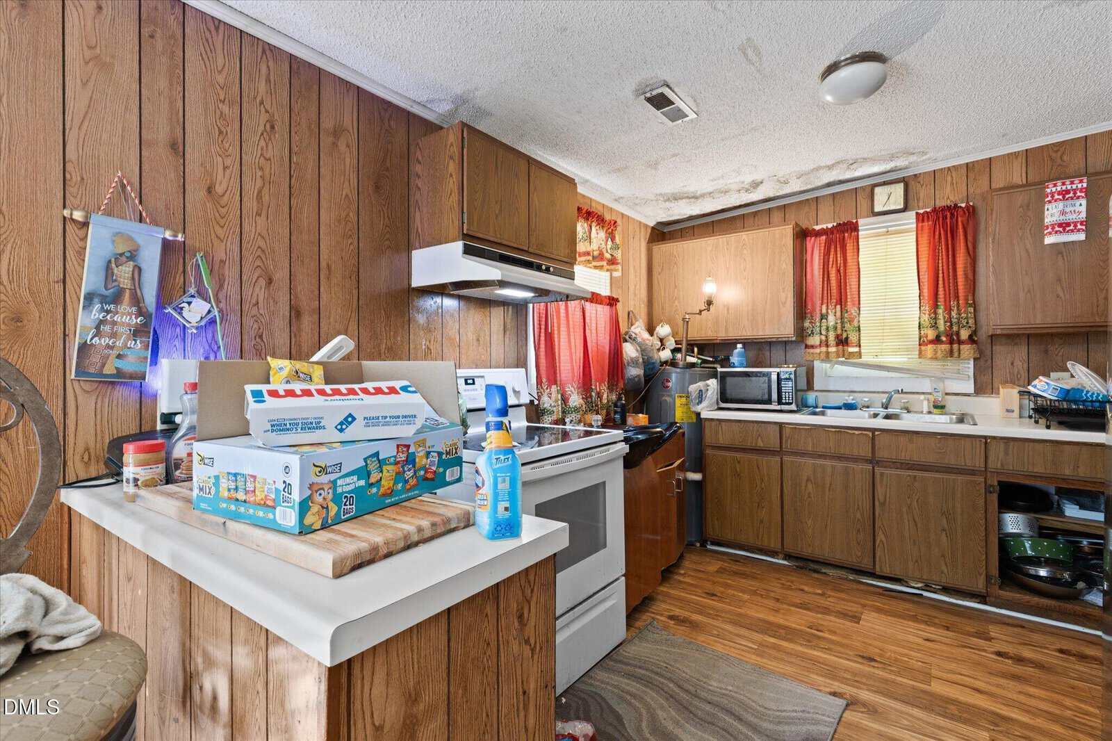 A small kitchen with wood panel walls, cluttered counters, red curtains, and various appliances. Board games and cleaning supplies sit on the counter near a stove and a sink filled with dishes.