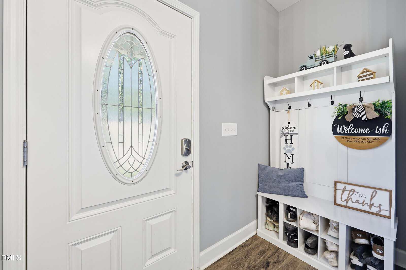 A white front door with an oval glass window is next to a white entryway bench with shoe cubbies, hooks, pillows, and welcome decor. The walls are gray and the floor is wood.