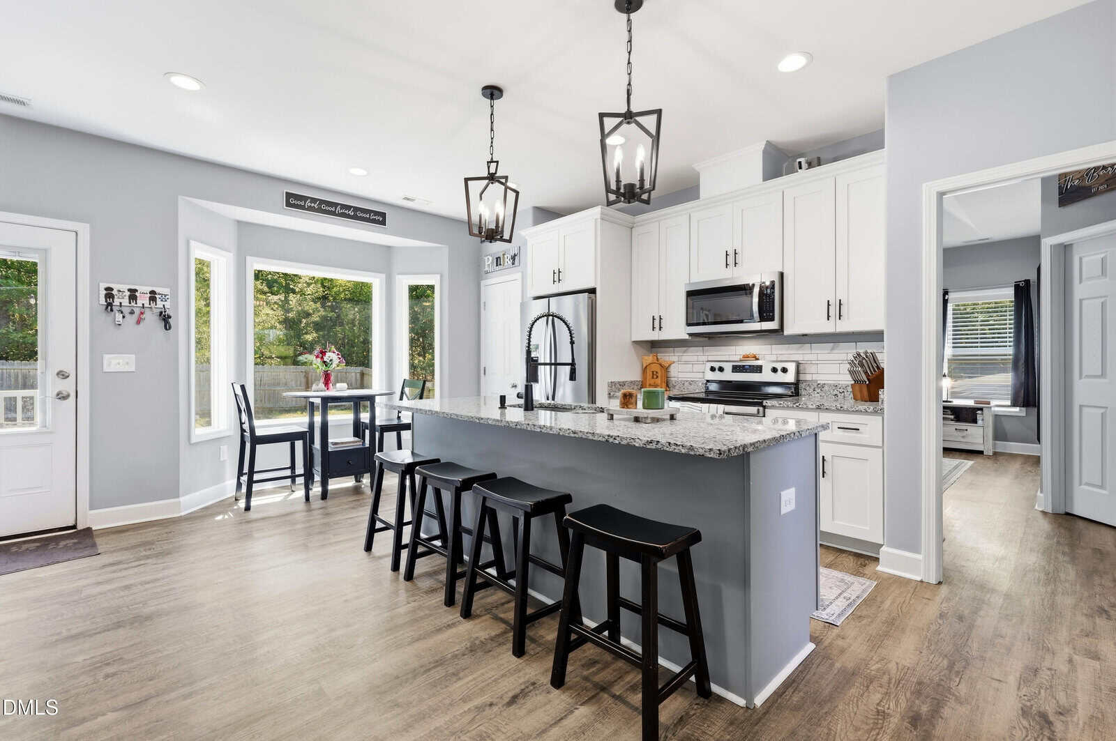 Modern kitchen with white cabinets, granite island, three black stools, stainless steel appliances, and pendant lights. Dining nook with table by large windows, and door leading outside; adjoining bedroom visible through open doorway.
