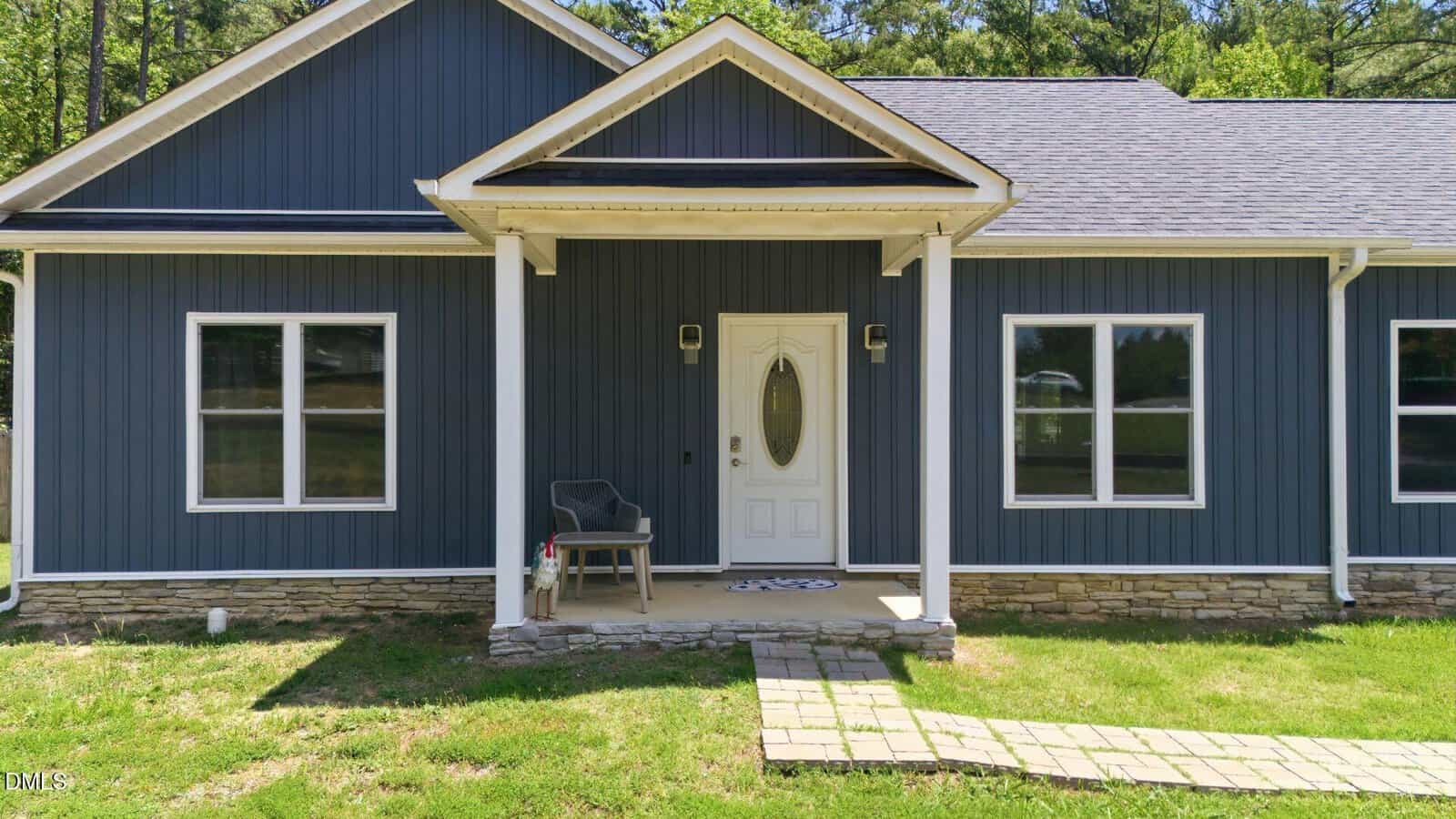 A blue house with white trim, stone accents, and a small front porch featuring a chair and doormat. A stone path leads to the white front door, with green grass and trees in the background.