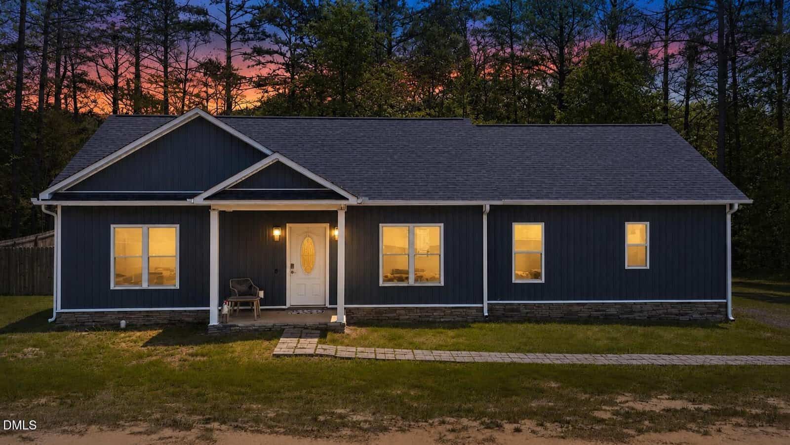 A single-story dark blue house with white trim is shown at dusk at 106 Old NC 75 N in Stem. The porch light is on, illuminating the front door and a chair nearby. Four windows are visible, and there are trees in the background against a colorful sky.