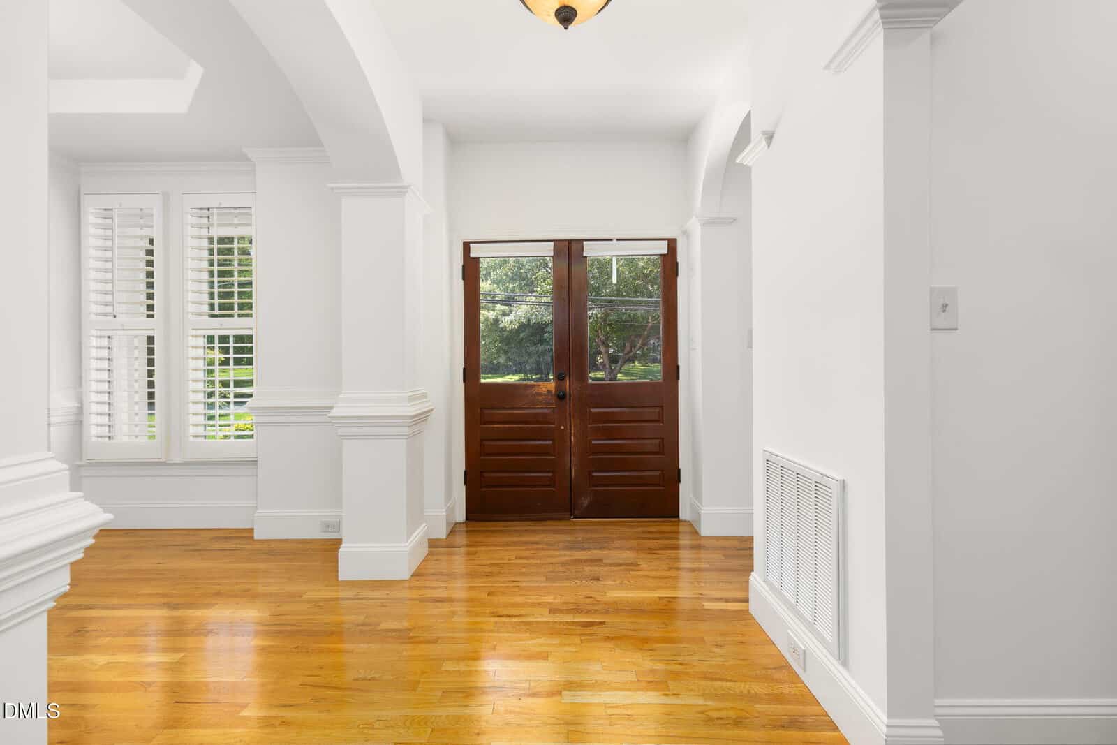 Bright entryway with hardwood floors, white walls, large windows with shutters on the left, and double wooden front doors with glass panels, allowing natural light to fill the space.