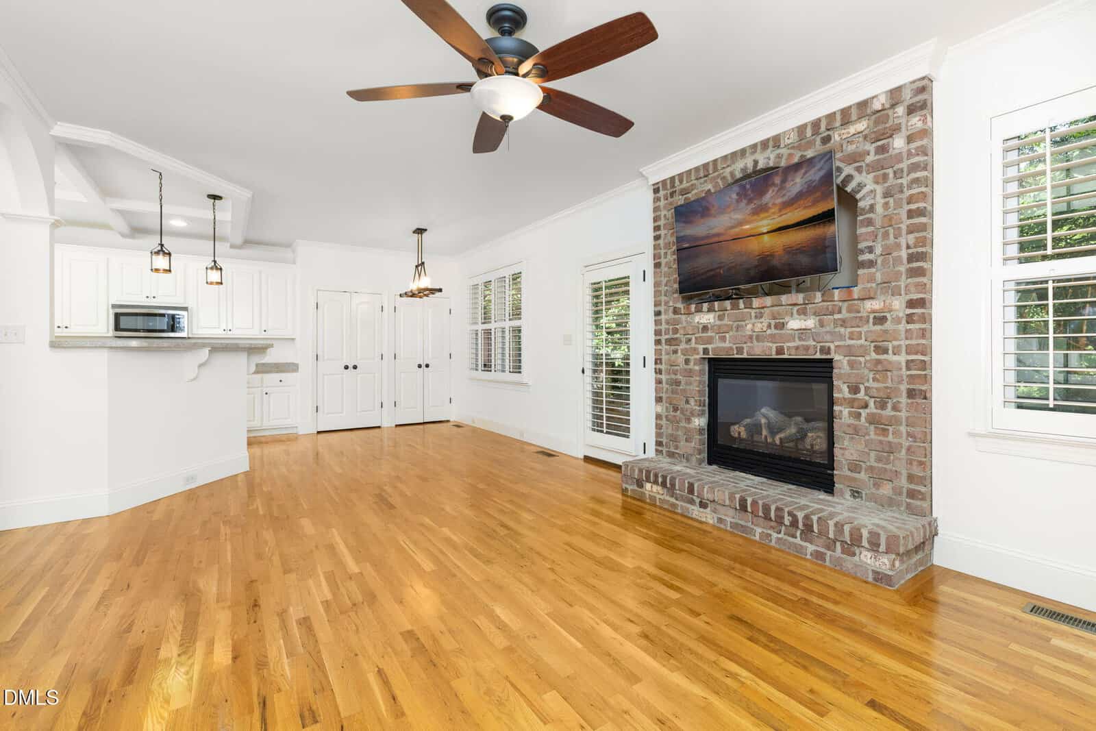 Bright living space with hardwood floors, brick fireplace, mounted TV, white walls, ceiling fan, and large windows. Kitchen with white cabinets and pendant lights is visible in the background.