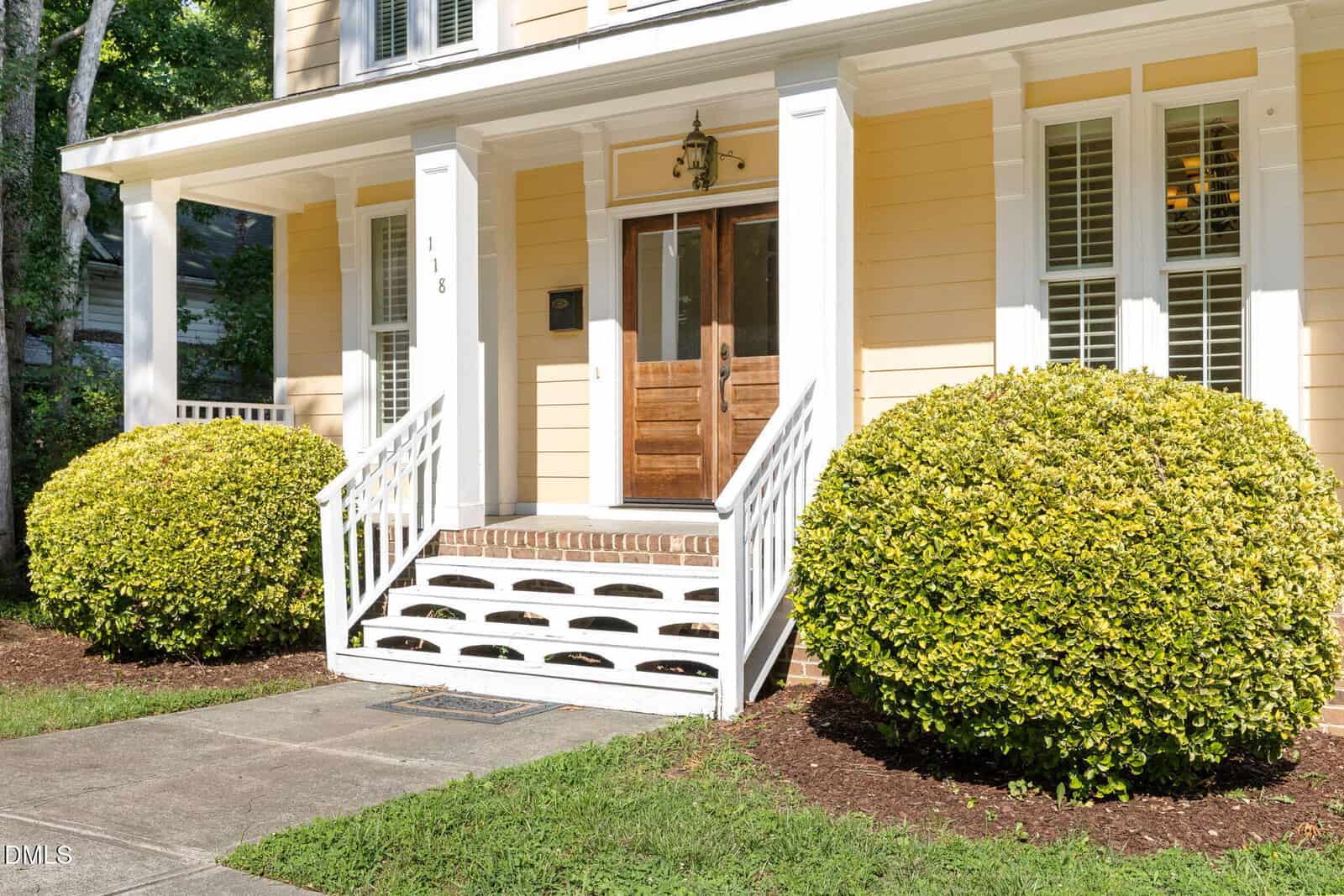 Yellow house with white trim, a wooden front door, white porch railing, two large green bushes on either side of the steps, and sunlight casting shadows across the entrance and walkway.