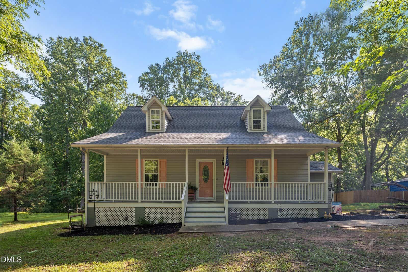 A light gray house with a covered front porch, two dormer windows, and an American flag. Surrounded by green trees and a lawn under a blue sky with scattered clouds.