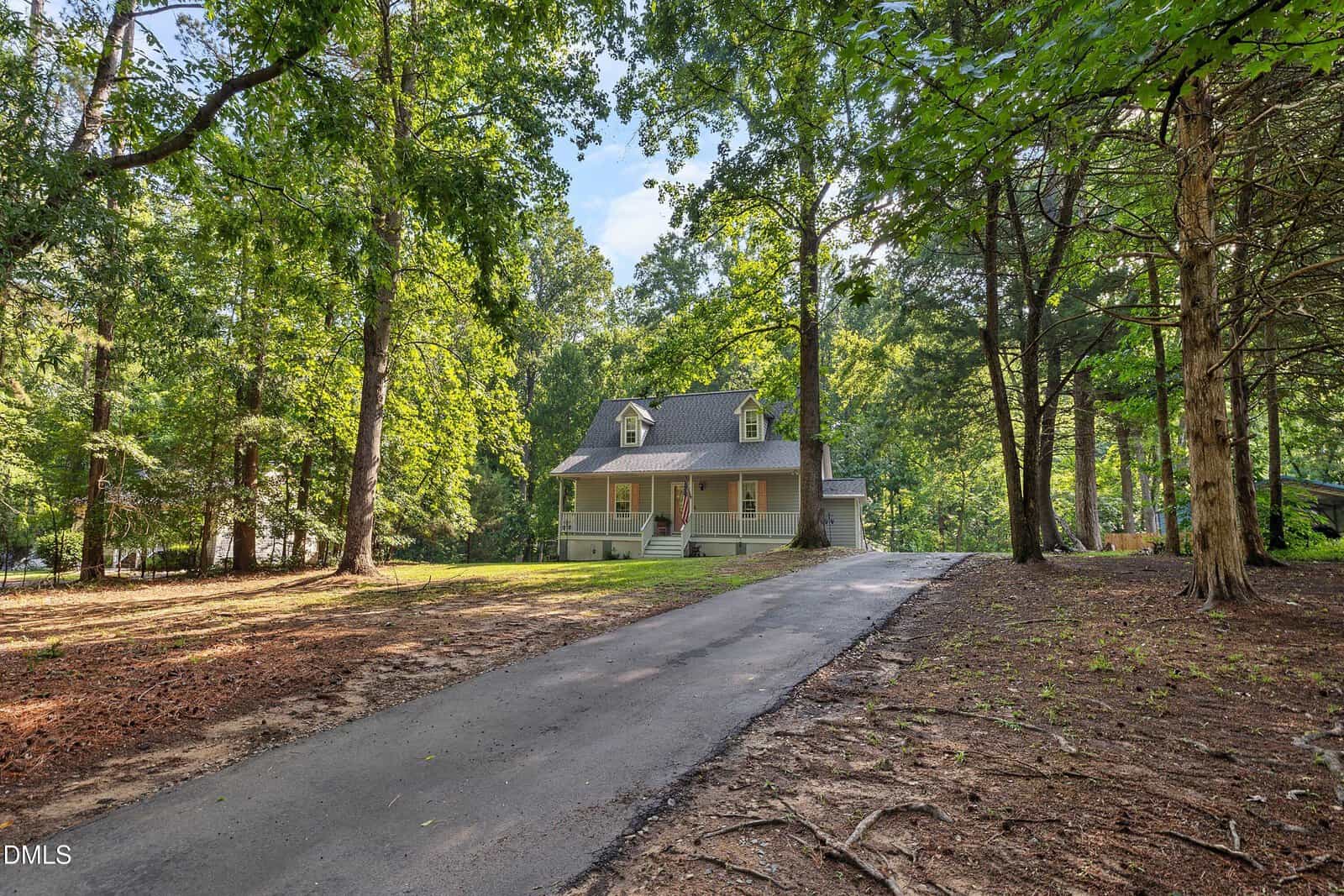 A gray house with a front porch sits at the end of a paved driveway, surrounded by tall trees and a wooded yard on a sunny day.
