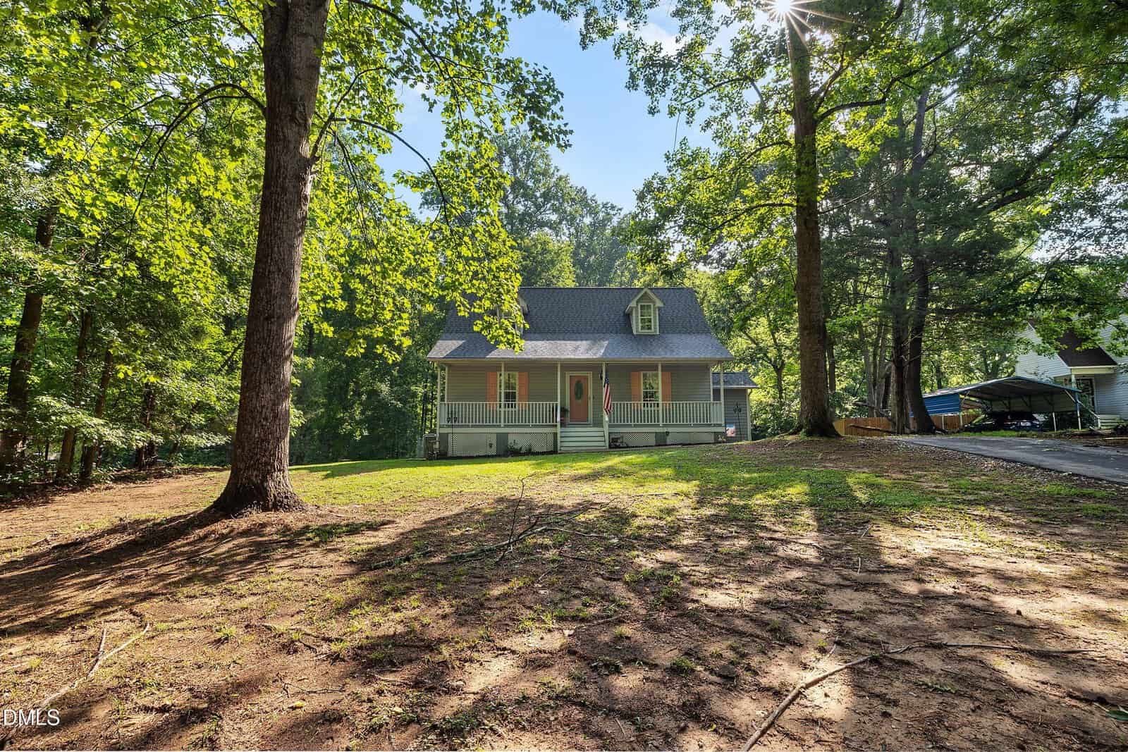 A light gray house at 112 Waiters Way in Youngsville, with a covered front porch, sits among tall trees, sunlight filtering through the leaves onto a spacious, partially shaded yard.