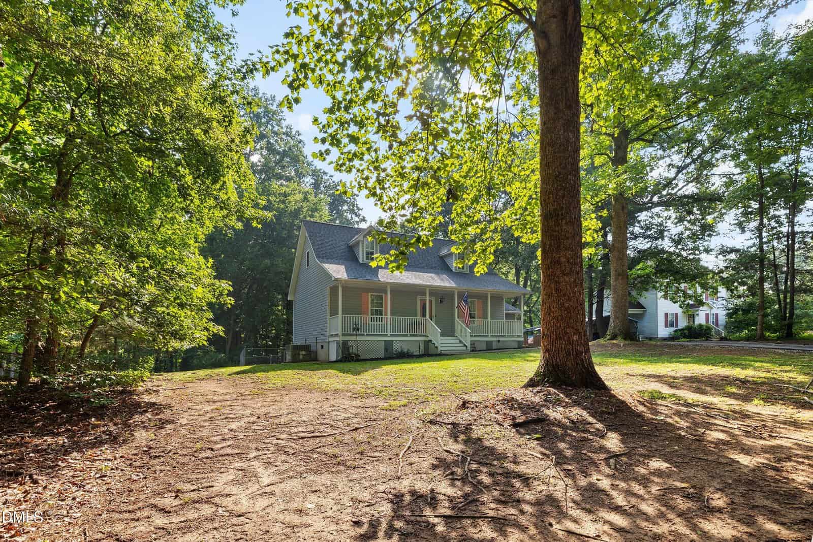 A light gray house with a front porch sits among tall trees, with sunlight filtering through the leaves onto a grassy, partially bare yard.