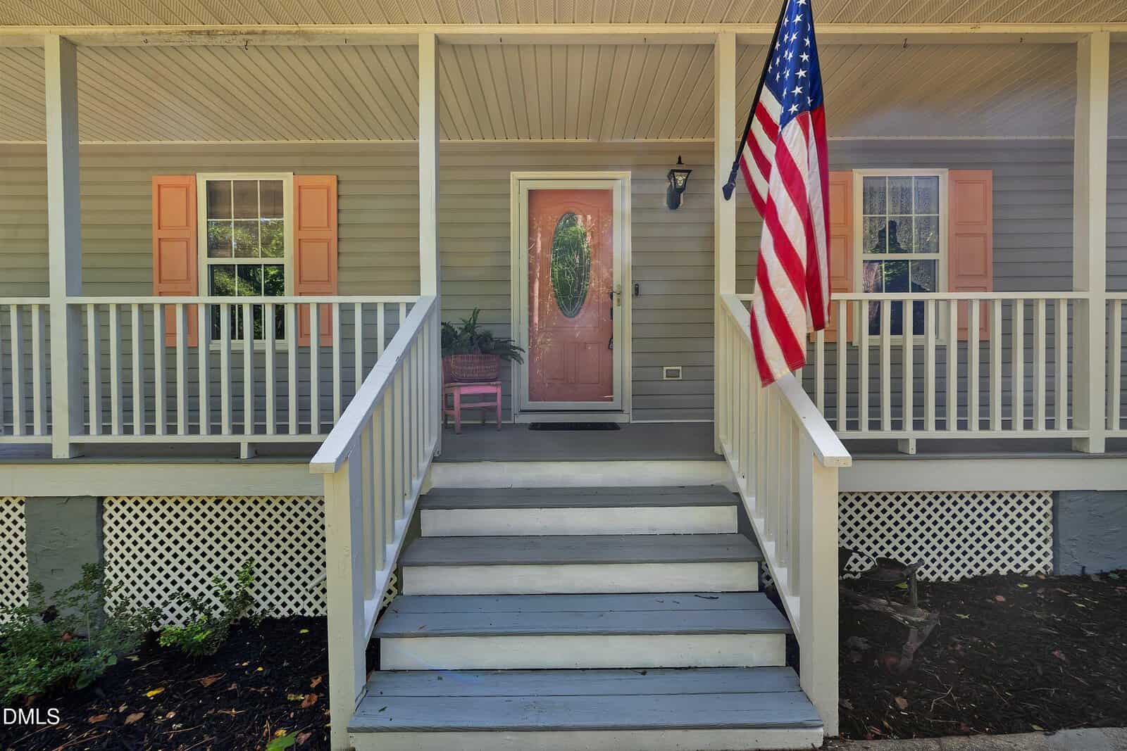 Front view of a house with a covered porch, American flag, light gray steps, peach front door, white railing, lattice trim, and two windows with peach shutters. Shrubs and mulch line the ground.