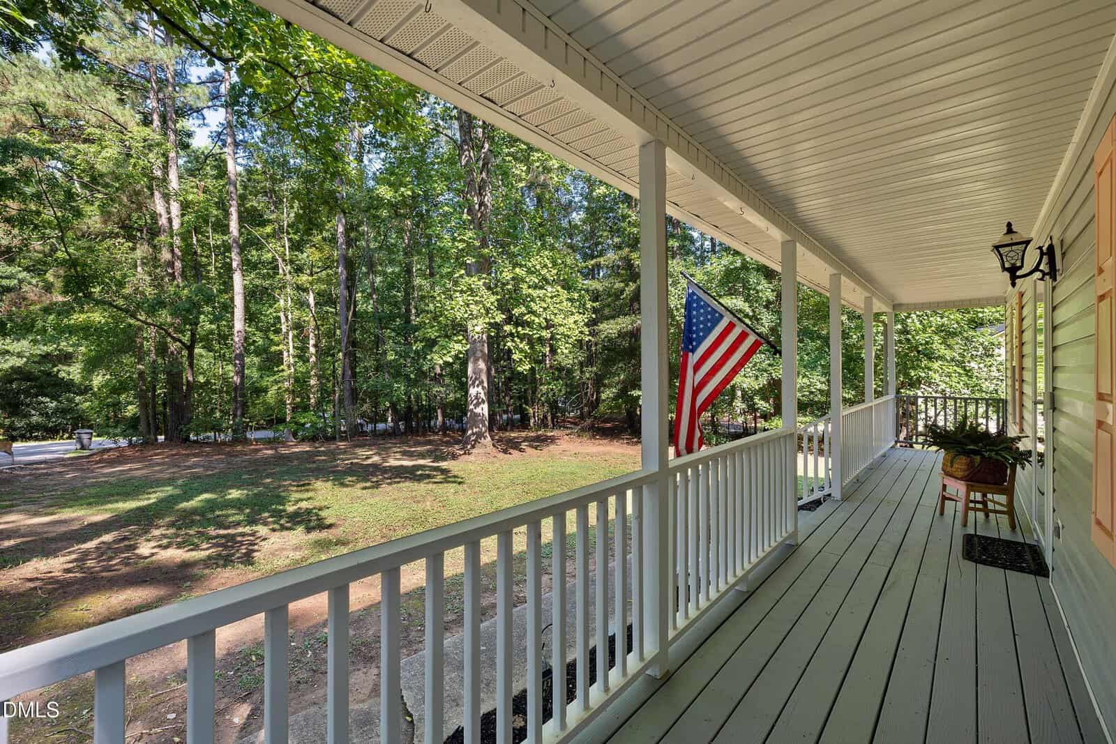 A spacious front porch with white railings and an American flag overlooks a large, wooded yard with tall trees and green grass. A potted plant sits near the porch door.