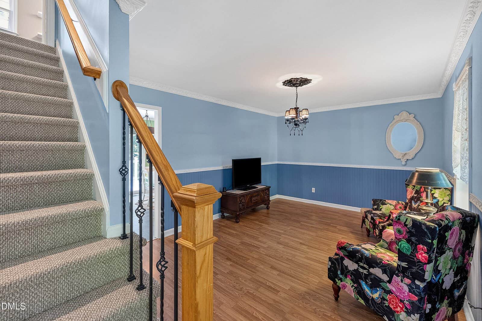 Living room with blue walls, wood floor, floral armchair, TV on a wooden stand, decorative mirror, chandelier, and carpeted stairs with a wooden handrail.