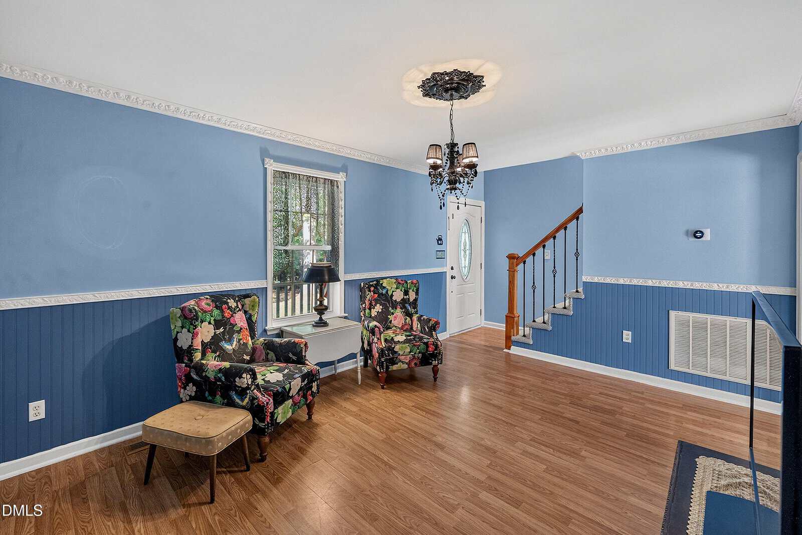 Living room with blue walls, wood floor, floral armchairs, a small beige bench, a window with curtains, a chandelier, and stairs beside a white front door.