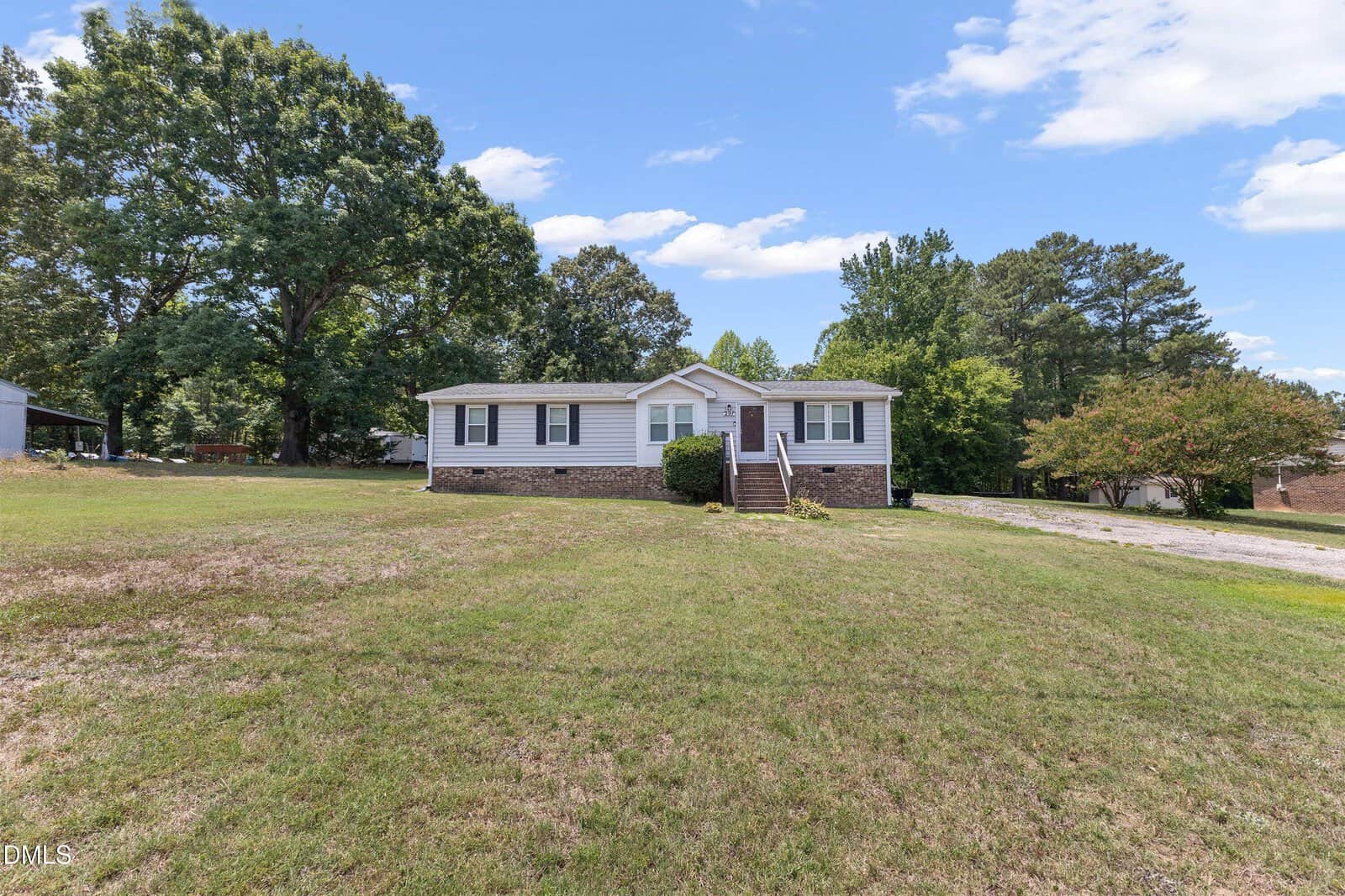Single-story house with white siding and brick foundation, front steps leading to a small porch, large grassy yard, trees in the background, and a clear blue sky with scattered clouds.