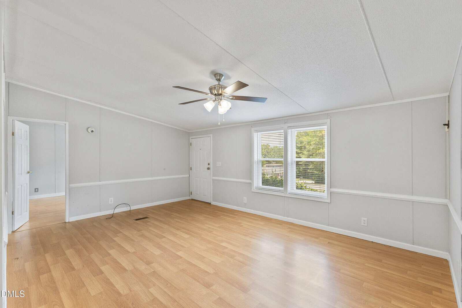 Empty room with light wood flooring, gray walls, white trim, a ceiling fan with lights, and two large windows letting in natural light. An open doorway leads to another room with the same flooring.